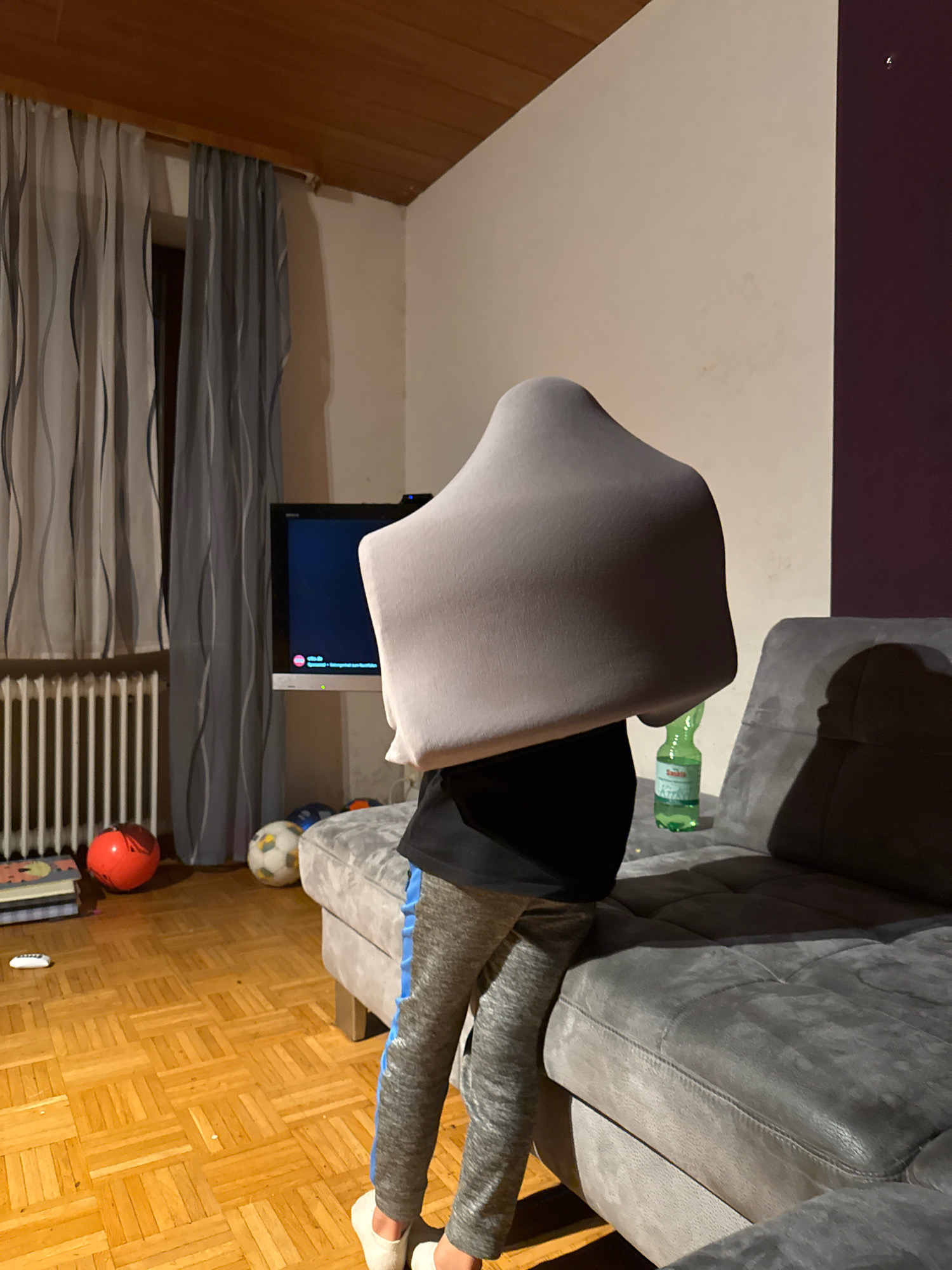 A person, whose face is obscured by a grey blanket draped over their head and upper body, stands in a living room. The person is wearing grey sweatpants with a blue stripe and white socks. They are positioned between a television set on a stand and a large grey sectional sofa. A green bottle of water is on the sofa next to the person. The room has light-colored walls, dark wood-paneled ceiling, and a light wood parquet floor. In the background, there are grey curtains, a radiator, and several soccer balls on the floor. The overall scene suggests a casual, indoor activity, possibly playtime or relaxation.