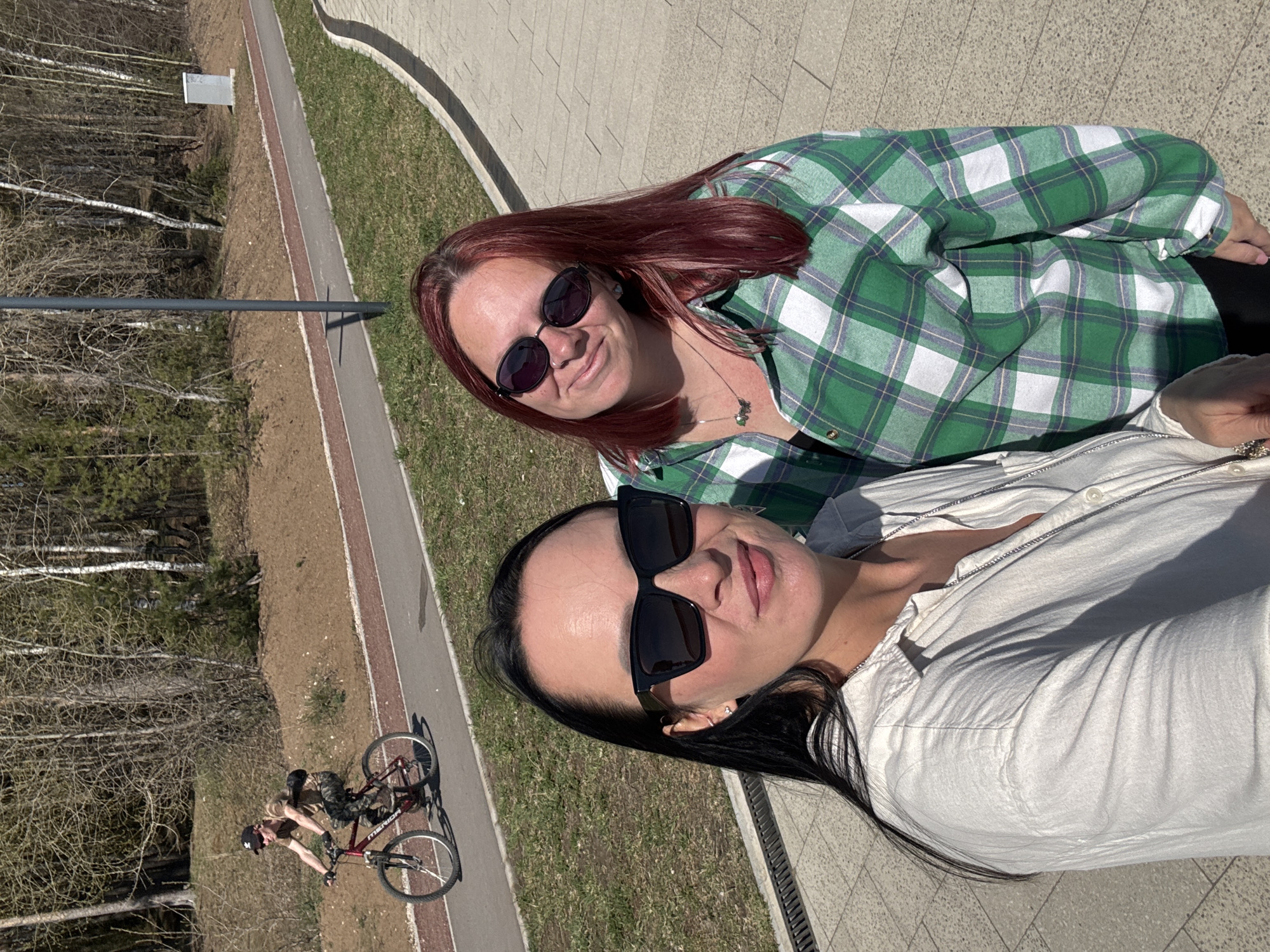 Two women are posing for a selfie outdoors. The woman on the left has long, dark hair and is wearing a white shirt and large black sunglasses. The woman on the right has red hair and is wearing a green and white plaid shirt with black sunglasses. They appear to be standing on a paved surface next to a grassy area. In the background, there is a bicycle path with a person riding a bicycle. The path runs alongside a forest with bare trees. A concrete wall or barrier is visible in the upper right corner. Based on the geolocation, the photo was taken in the city of Chaikovsky, Perm Krai, Russia.