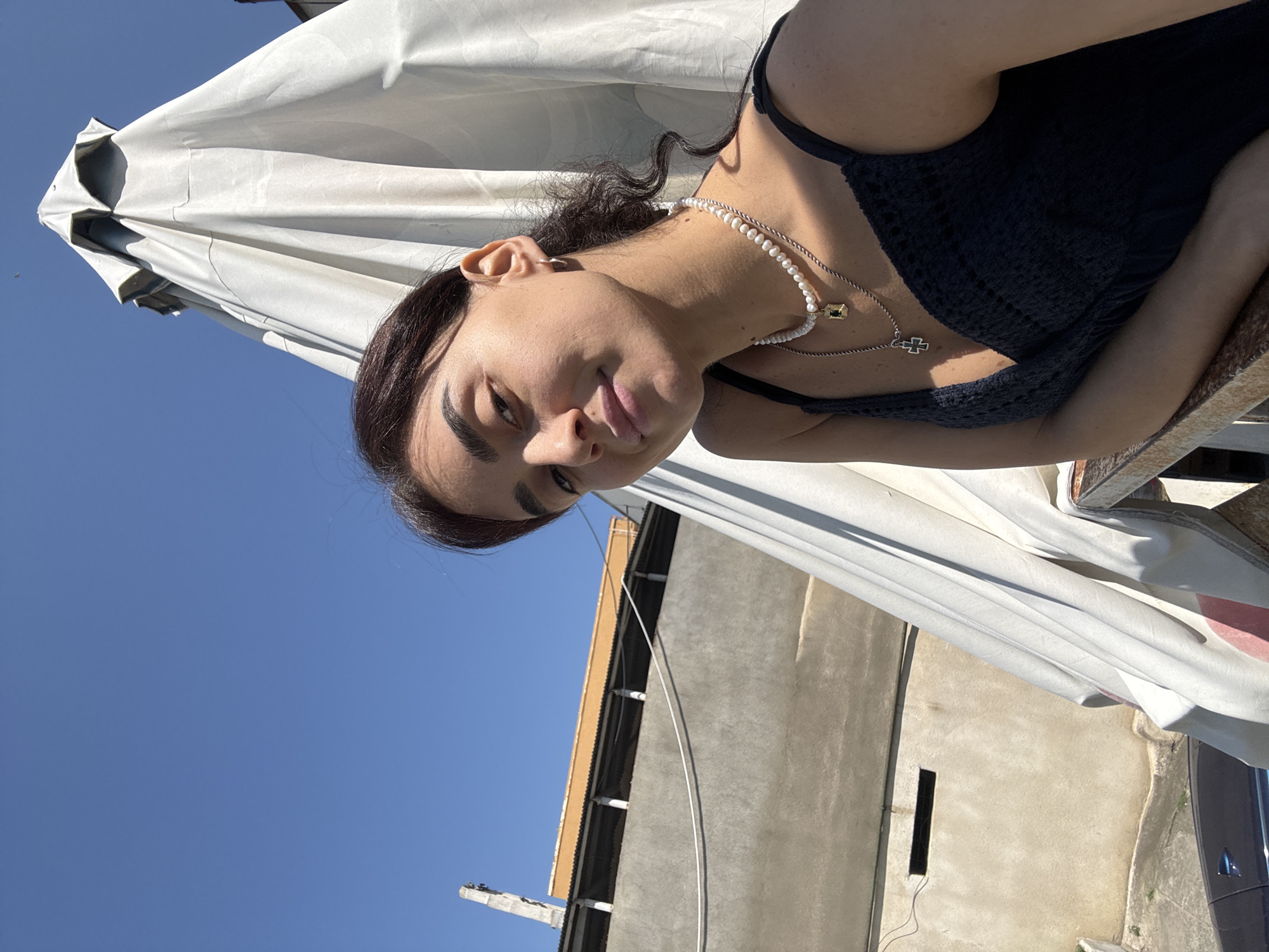 The image is a selfie taken outdoors on a sunny day in Tbilisi, Georgia. A young woman with dark hair pulled back is looking towards the camera, her face illuminated by sunlight. She is wearing a dark blue dress with thin straps and a crochet detail, and layered necklaces, including a pearl one and one with a cross pendant. Behind her, a large, white umbrella is partially covering the scene. In the background, a concrete building with an orange-colored top section and a corrugated roof is visible, along with a clear blue sky. To the left, a portion of a dark-colored car is visible. The environment suggests an urban setting, possibly a rooftop or balcony.