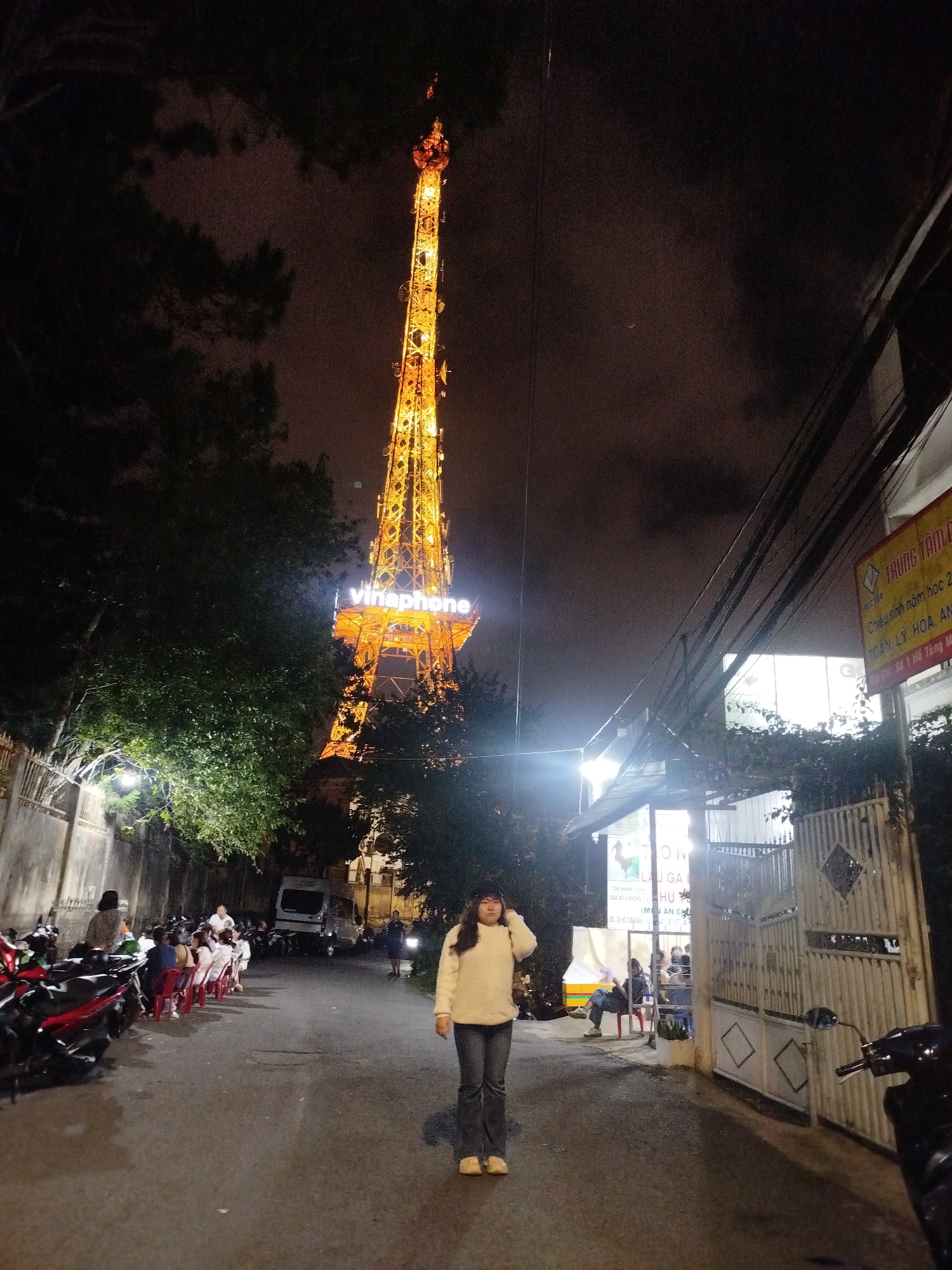 A nighttime street scene in Da Lat, Vietnam, features a towering, illuminated telecommunications tower as the dominant subject. The tower, adorned with the "vinaphone" logo, is brightly lit in orange and yellow hues against a dark, cloudy sky, resembling the Eiffel Tower. In the foreground, a young woman stands facing the camera, wearing a cream-colored sweater and jeans. She appears relaxed, with one hand near her head.
The street is lined with parked motorcycles on the left and a building with a white fence on the right. A white van is parked further down the street. Several people are seated at tables on the left side of the road, suggesting an outdoor dining or gathering area. The building on the right has a sign with Vietnamese text, reading "TRUNG TAM CHIEU SINH NAM HOC 2 TOAN LY HOA AN" which translates to "Center for Enrollment Academic Year 2: Math, Physics, Chemistry, Biology". There are also overhead wires and streetlights, indicating an urban environment. The overall atmosphere is one of a lively evening gathering under the glow of the prominent tower.