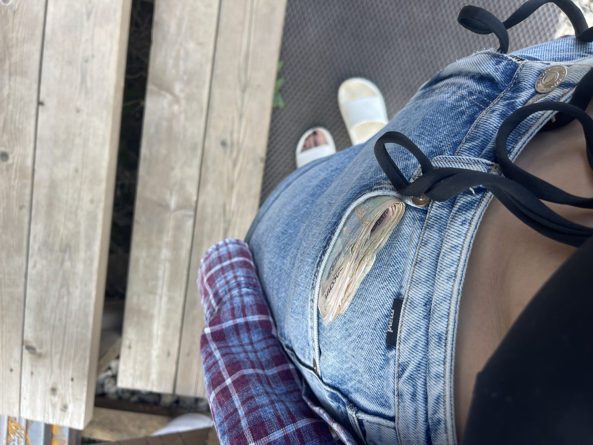 The image shows a person sitting, wearing blue jeans with a wad of Russian rubles sticking out of the pocket. They are also wearing white sandals and a red and blue plaid shirt is visible to the left. The background shows a wooden plank surface. Based on the city specified, the image location is Мосметаллоконструкция Онт, Russia.