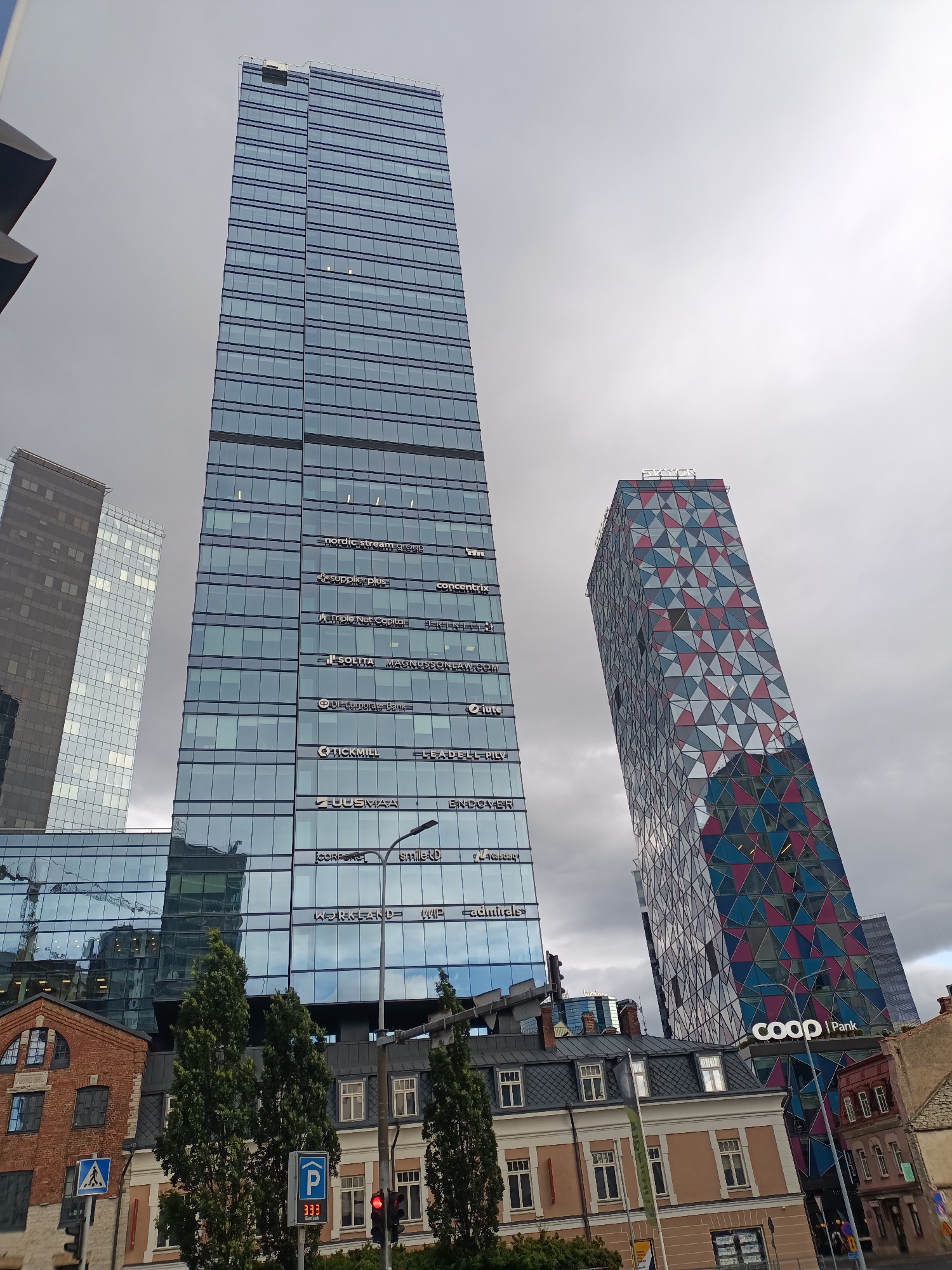 The image shows a street scene in Tallinn, Estonia, focusing on modern skyscrapers and older buildings. Dominating the view is the large, glass-facaded SEB building, with the names of various companies visible on its exterior, such as "nordic stream group," "supplierPlus," and "SEB Corporate Bank." To its right, a smaller, brightly colored skyscraper, featuring a geometric pattern of red, blue, and gray triangles, stands at an angle. The "coop | pank" logo is clearly visible on its lower section. In the foreground, a brick building and a row of older, two- and three-story houses with pitched roofs line the street. A pedestrian crossing sign, a parking sign with the number "333," and a traffic light are also present. The sky is overcast, suggesting a cloudy day. No people are prominently featured in the image.