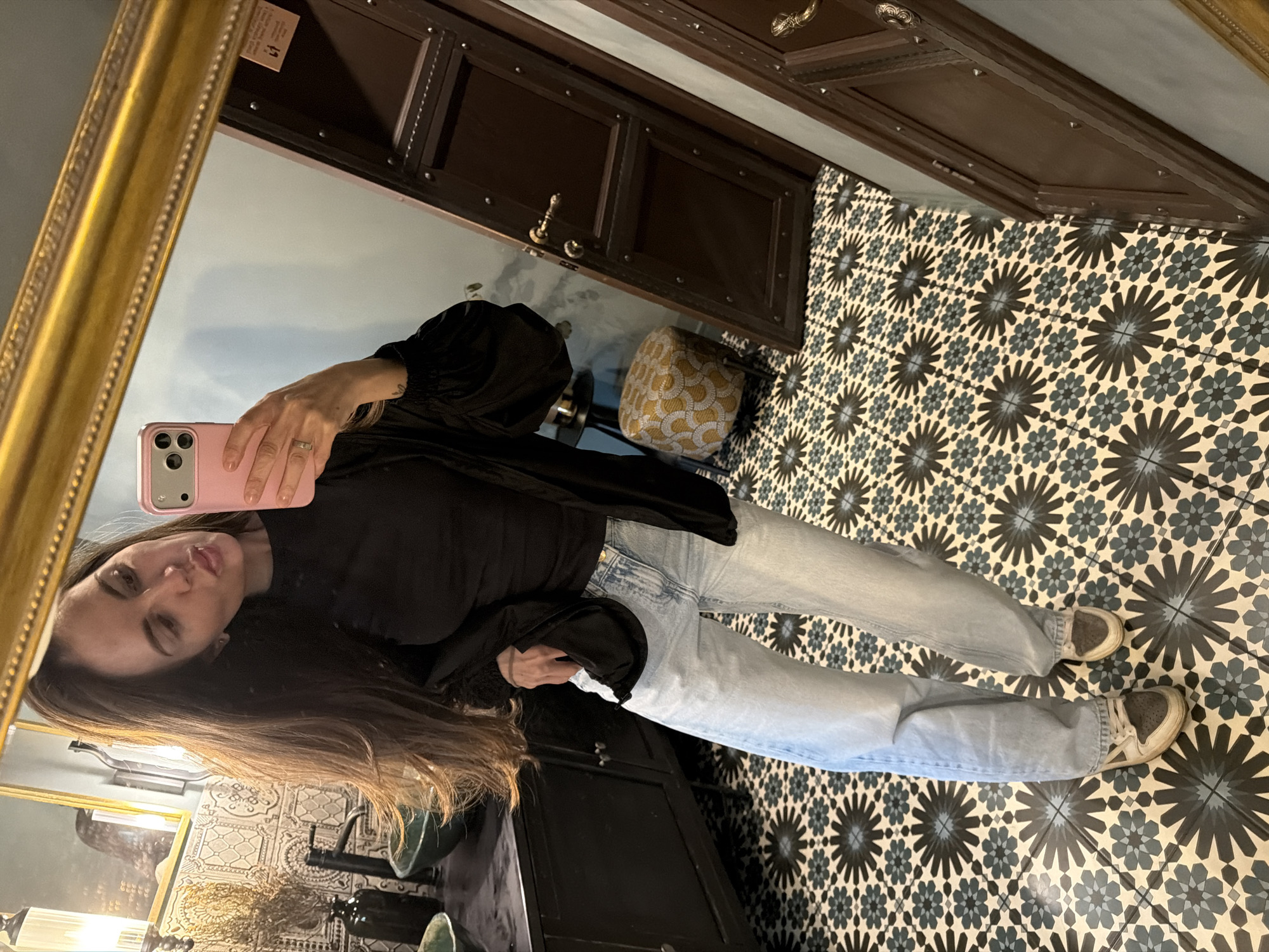A woman with long brown hair is taking a selfie in a mirror. She is wearing a black shirt, black jacket, and light blue jeans. Her feet are visible, wearing white shoes. The mirror has a gold frame. In the reflection, there is a patterned stool and patterned tile floor. The vanity is dark wood and has a dark wooden door with dark bronze hinges. The bathroom also contains bronze fixtures. The location is Krasnoyarsk, Russia.