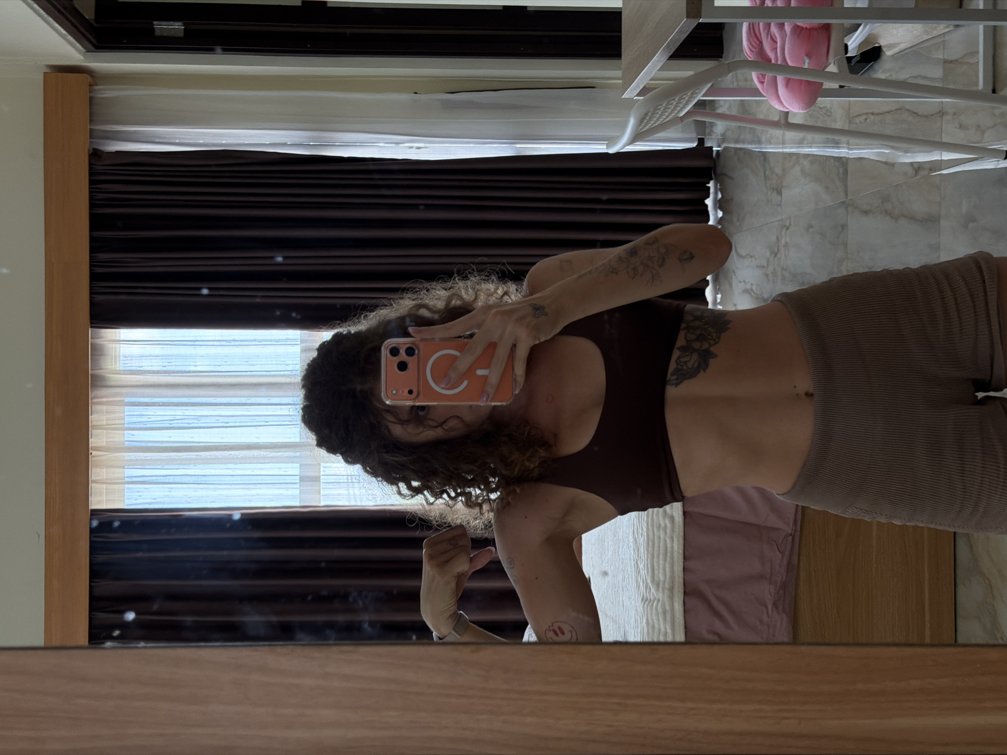 An eye-level mirror selfie captures a person in an indoor setting, likely a bedroom, in North Kuta, Indonesia, during the daytime. The person, who has long curly dark hair and is wearing a dark brown sports bra or crop top and light brown ribbed shorts, is reflected in a wooden-framed mirror. They are holding an orange smartphone with a white MagSafe ring and faintly visible text "C-" with a lightning bolt symbol and "12 PRO MAX" on the back, indicating the phone model. The person's left arm is slightly flexed, revealing tattoos including floral designs on their left shoulder/arm and torso (left ribcage area), and a small melting smiley-face tattoo on their left forearm.
The background reflected in the mirror shows a room with light-colored tiled flooring and a window partially covered by dark brown horizontal blinds over sheer white blinds. A bed with a light pink sheet and a white textured blanket is visible to the right. Other reflected objects include a light-colored metal chair or stool, a pink fluffy item (possibly a cushion or garment), and a black shoe on the floor, all positioned towards the right side of the frame. The lighting suggests natural daylight entering the room. The person's expression is neutral, focused on taking the photograph.