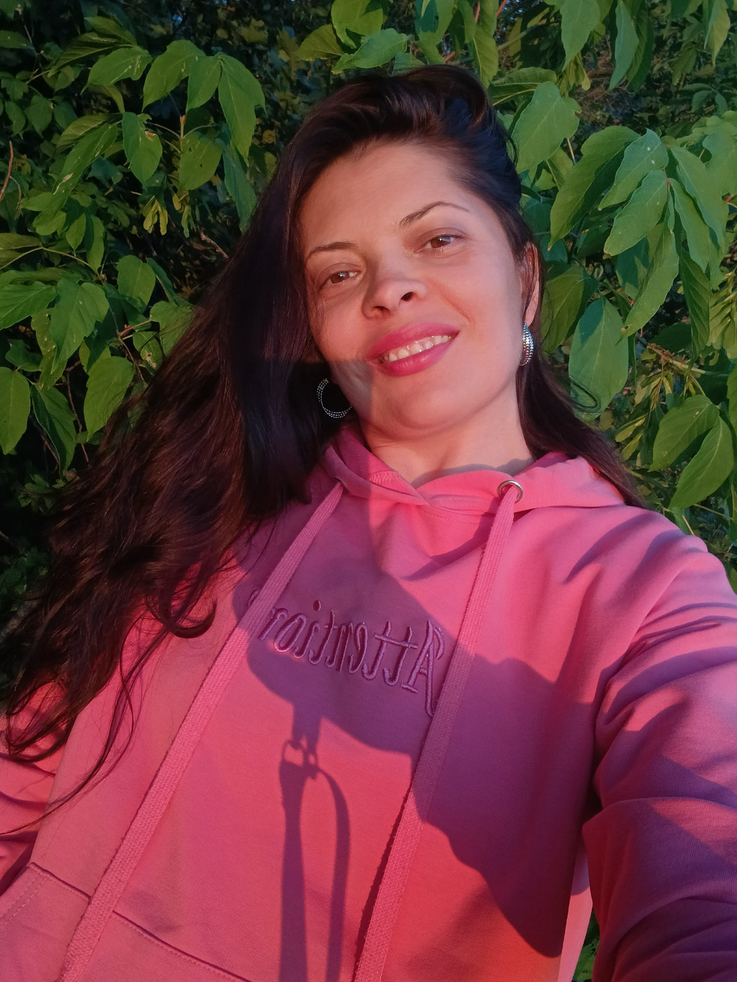 A woman with long, dark hair and wearing a pink hooded sweatshirt is pictured outdoors. She is smiling and looking towards the camera. The background consists of lush green foliage. The lighting suggests it is either early morning or late afternoon, with warm golden hour light illuminating the scene. The woman is wearing hoop earrings. The sweatshirt has embroidered text on the front, though the text is partially obscured and difficult to read. Based on the available information, it is not possible to determine the specific location in Saratov, Russia.