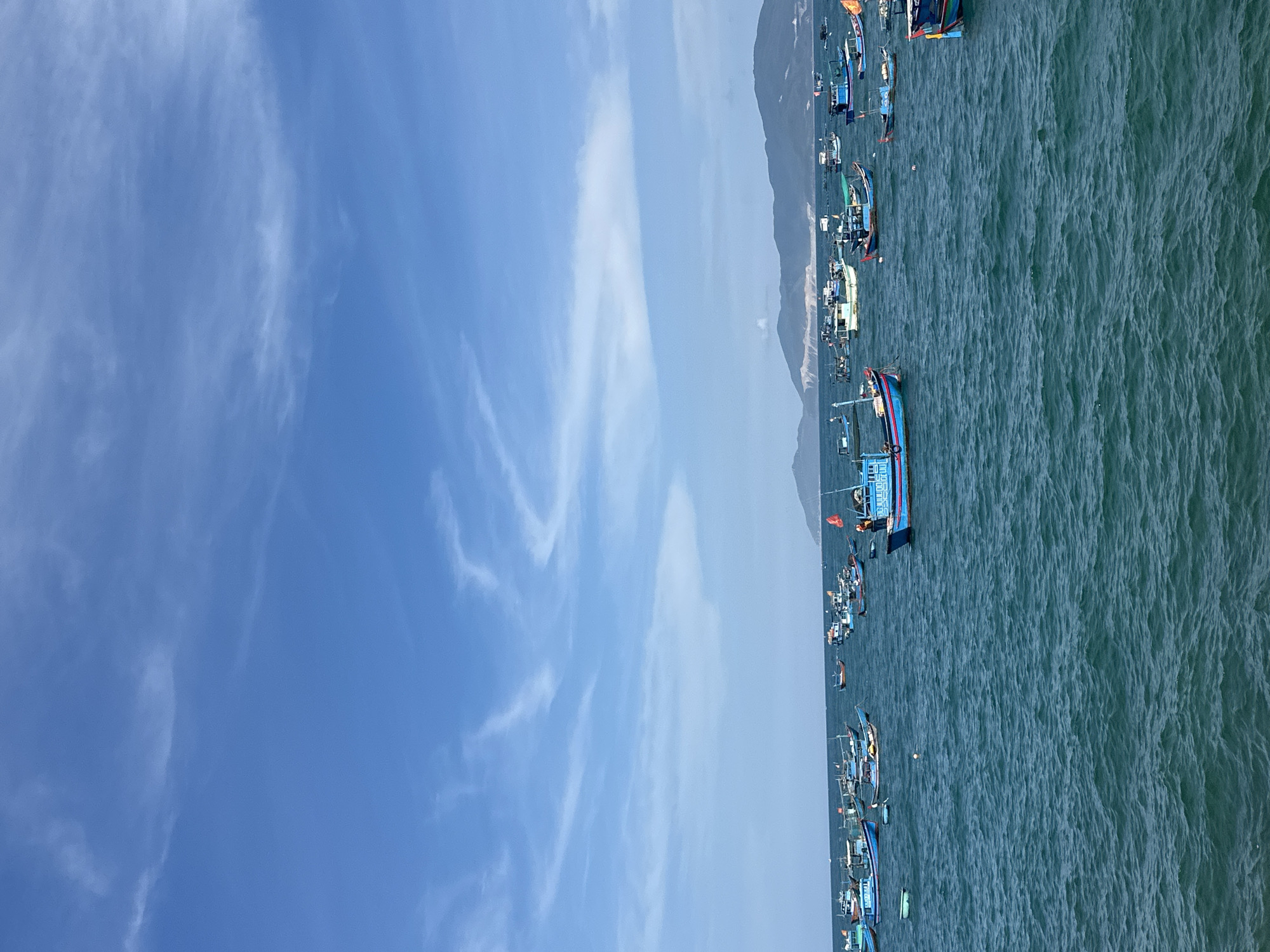 The image shows an aerial view of Nha Trang Bay near Bac Nha Trang Ward, Vietnam. The sky is a clear blue with scattered cirrus clouds. A mountainous coastline is visible in the distance. In the foreground is the turquoise water of the bay, populated with numerous fishing boats, both large and small.