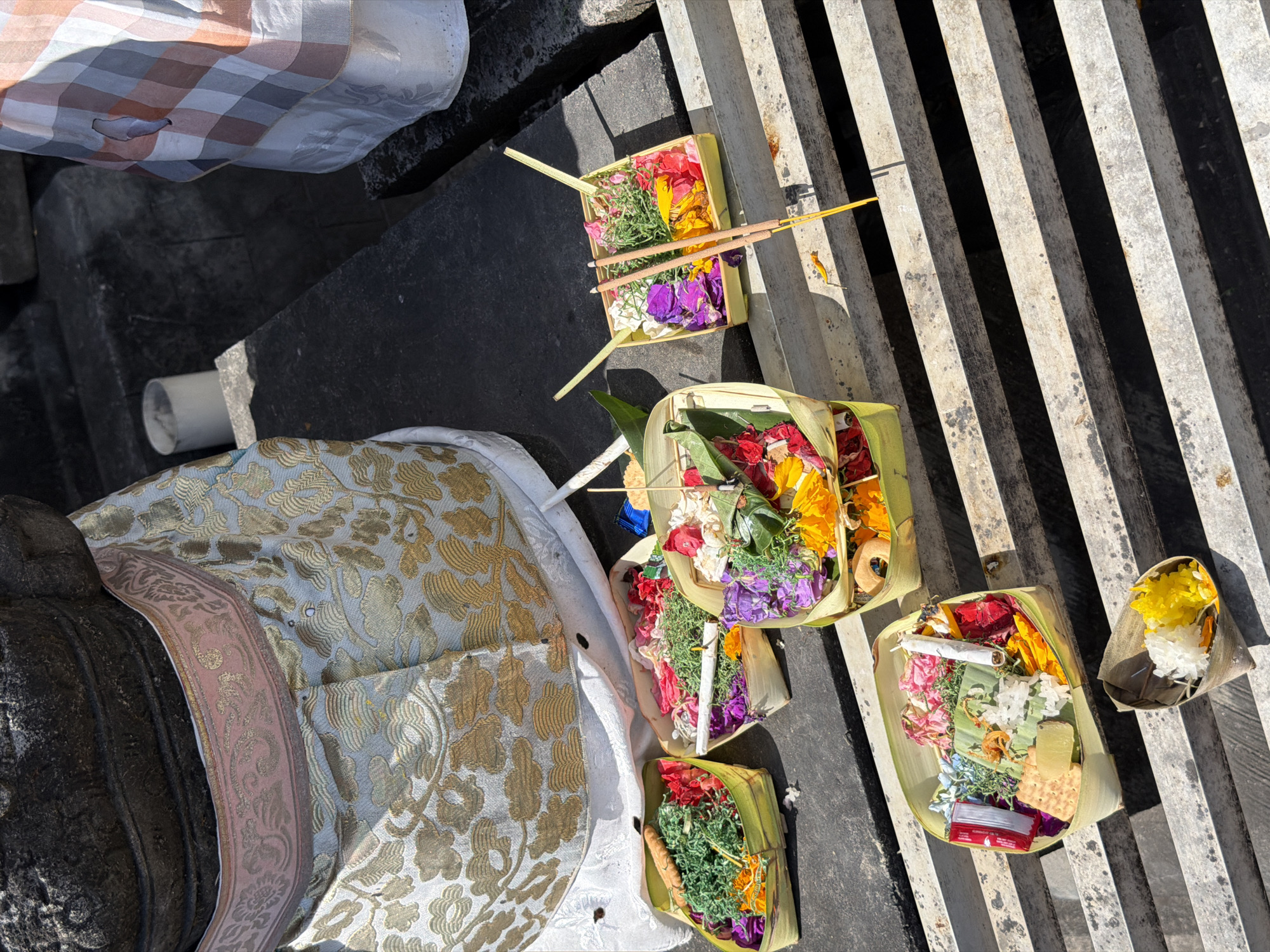 The image shows several offerings placed on a tiered, grate-like surface. The offerings are contained in woven palm leaf baskets and consist of colorful flower petals, green shredded material, incense sticks, and what appear to be small food items. In the background, a statue is partially visible, adorned with a patterned cloth. Another person is seen to the right, also wearing a patterned cloth. The environment suggests an outdoor setting, possibly a temple or religious site in Kuta, Indonesia, given the presence of offerings and the cultural attire. The lighting indicates it is daytime.