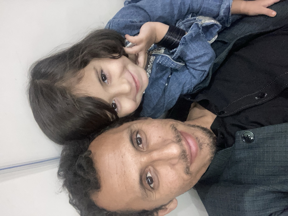 An adult male and a young female child are depicted in a close-up portrait against a plain, light-colored interior wall, specified as Quetta City Tehsil, Pakistan. The male, positioned on the left and partially in the foreground, has dark, curly hair, a mustache, and a short goatee. He is wearing a dark, possibly charcoal, collared jacket over a black collared shirt, and is looking directly at the camera with a subtle smile. The child, positioned to the right and slightly behind the male, has dark, shoulder-length hair and is wearing a blue denim jacket. Multiple dark bangles are visible on her left wrist. She is also looking towards the camera, displaying a broad smile, with her left hand positioned near her chin. The background is an undecorated light wall, featuring a faint vertical line in the upper left quadrant, consistent with an indoor setting.