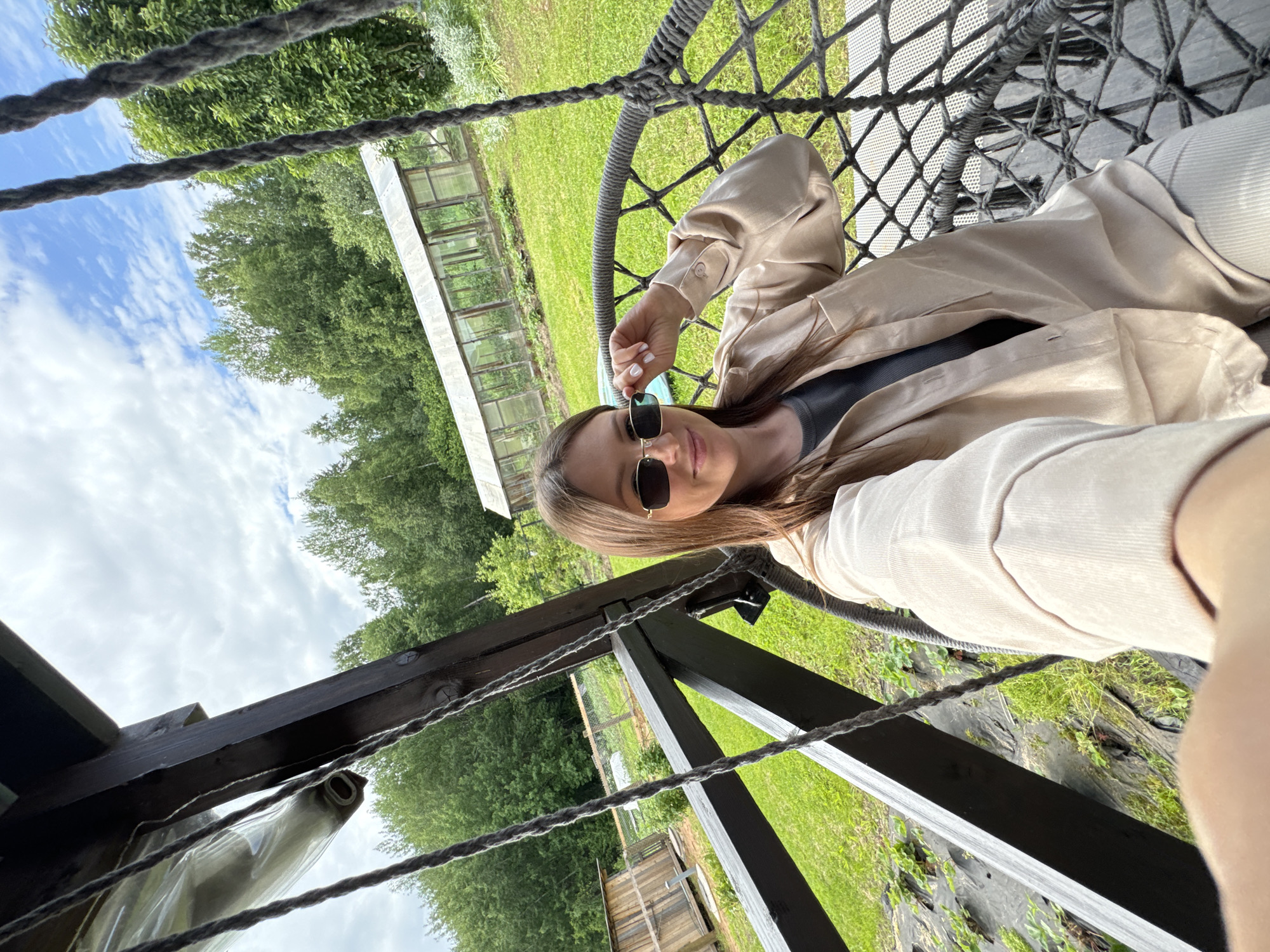 A young woman with long brown hair and sunglasses sits in a dark woven hanging chair on a wooden porch. She is wearing a light beige button-down shirt over a dark grey top and light beige pants. The woman is holding her sunglasses with her right hand and looking at the camera. In the background, there is a green lawn, trees, a greenhouse, and a wooden shed. The image appears to be taken in the rural area of Поназырево, Russia.