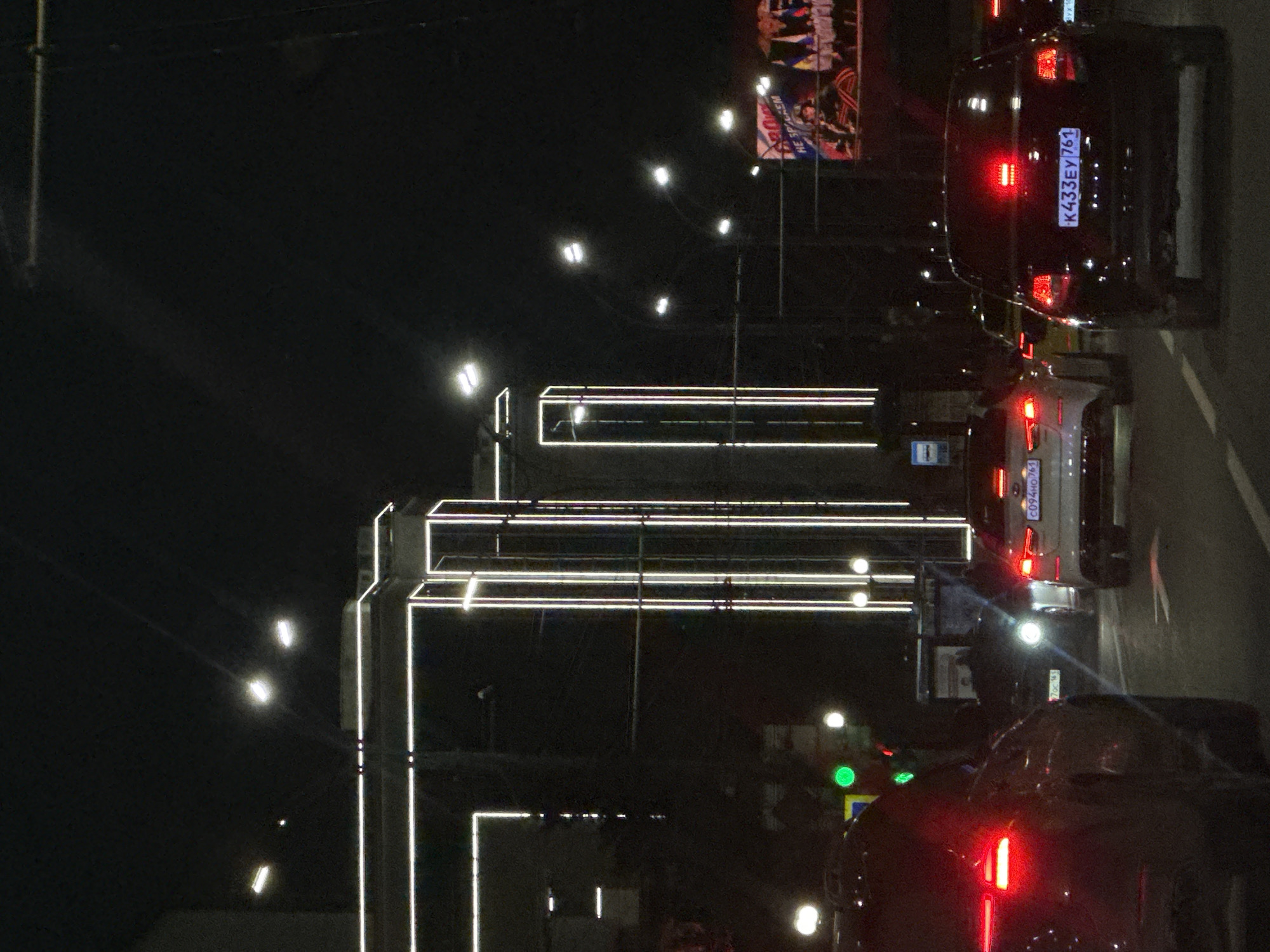 The image shows a nighttime scene in Rostov-on-Don, Russia, featuring cars on a street with illuminated buildings. The buildings have linear lights outlining their structure. Cars with illuminated tail lights are in the foreground and further down the street. Russian license plates are visible on the vehicles, with one clearly reading "K433EY761." Street lights and a billboard add to the urban environment. The scene is dark with artificial lights highlighting architectural elements and traffic.