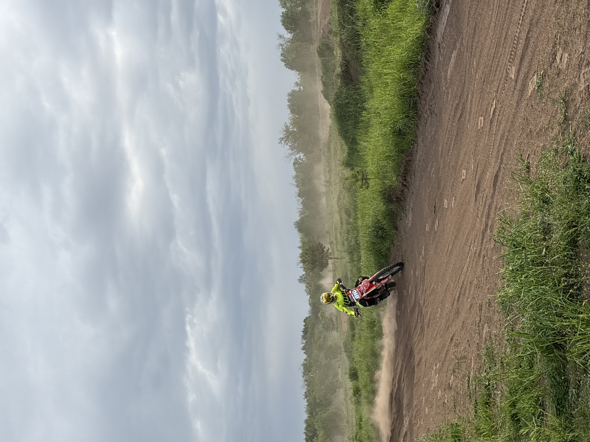 A motocross rider in neon yellow gear and a red motorcycle is maneuvering on a dirt track. The track is bordered by green vegetation. Dust is kicked up by the motorcycle's movement. The sky is overcast. It is not possible to determine whether the location is in Votkinsk, Russia from the image.