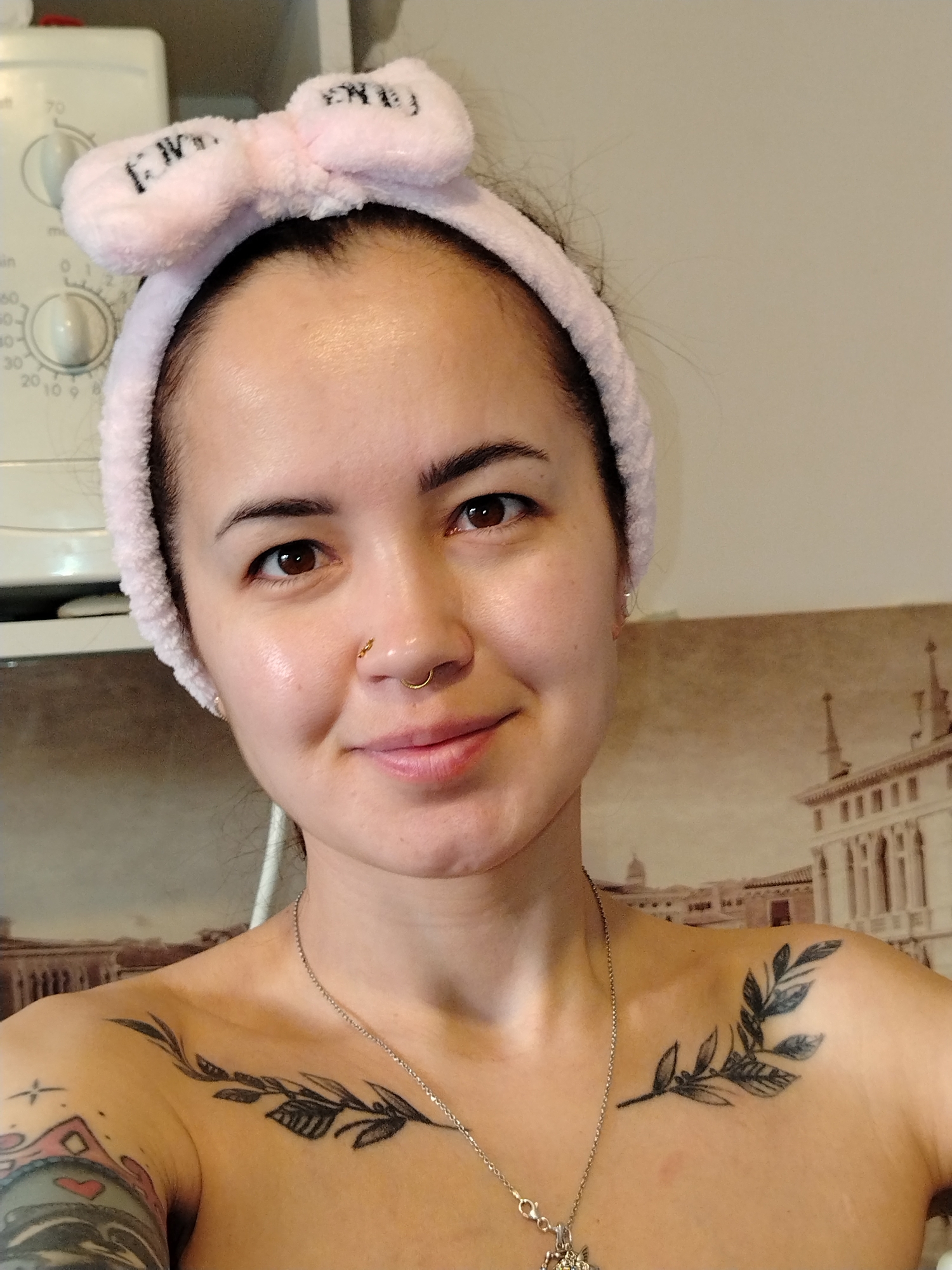 A selfie of a woman with fair skin, brown eyes, and dark eyebrows is presented. She is wearing a pink headband with a bow on top. A gold nose ring is visible in her left nostril. She is also wearing a thin silver chain with a pendant around her neck. Tattoos are visible on both shoulders. The left shoulder has a colored tattoo, and the right shoulder has a black-ink branch tattoo. In the background, there is a wall with a print of a city skyline, reminiscent of European architecture. The city skyline in the background appears to represent Venice, Italy, and is not located in Noviy, Russia. A white appliance, possibly a washing machine, is visible on the top left.