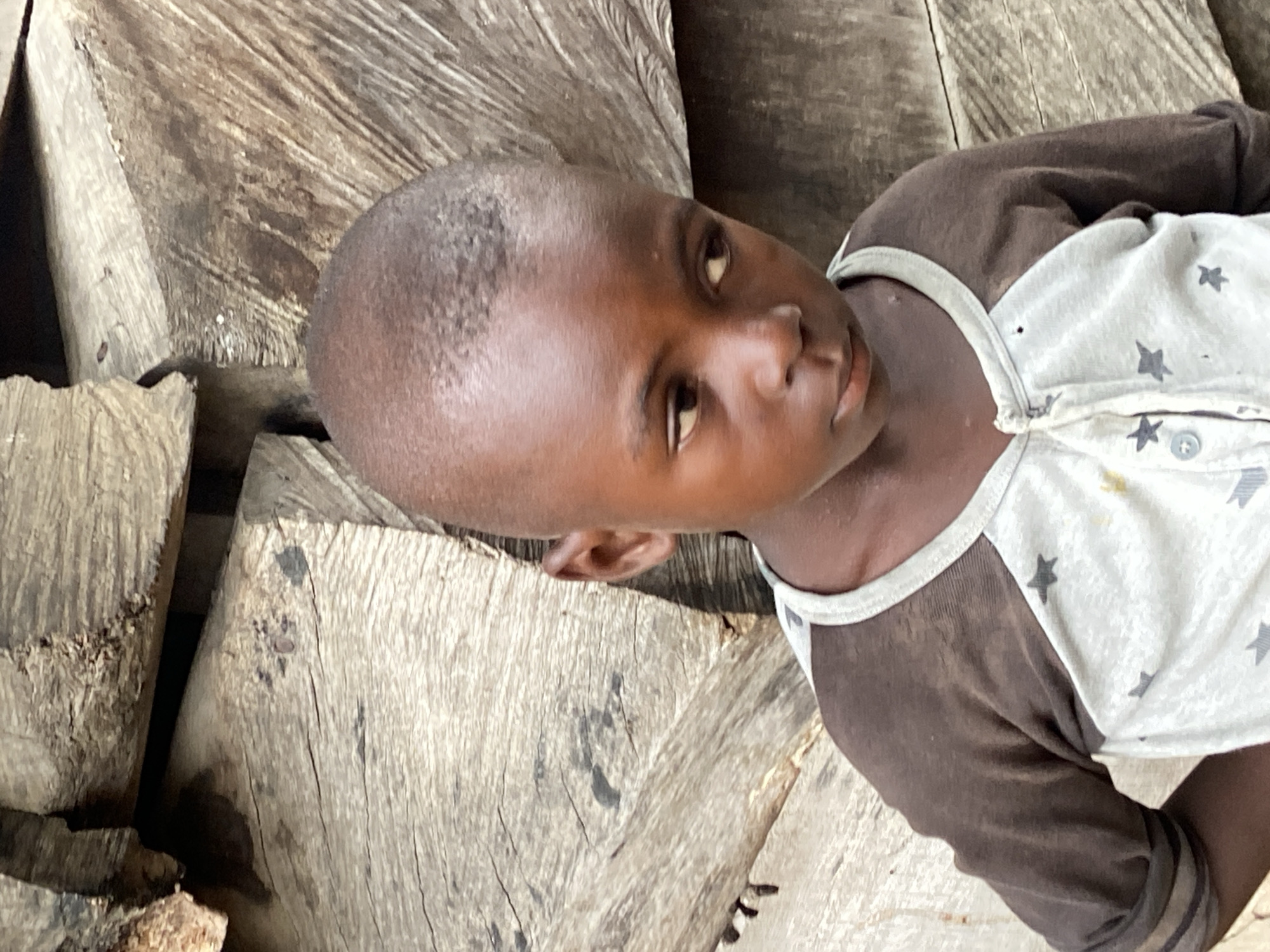 A child lies on rough, unfinished wooden planks outdoors. The child is Black with a shaved head and wears a brown and white star-patterned shirt. Based on the location provided, the image likely depicts Mararaba, Nigeria.