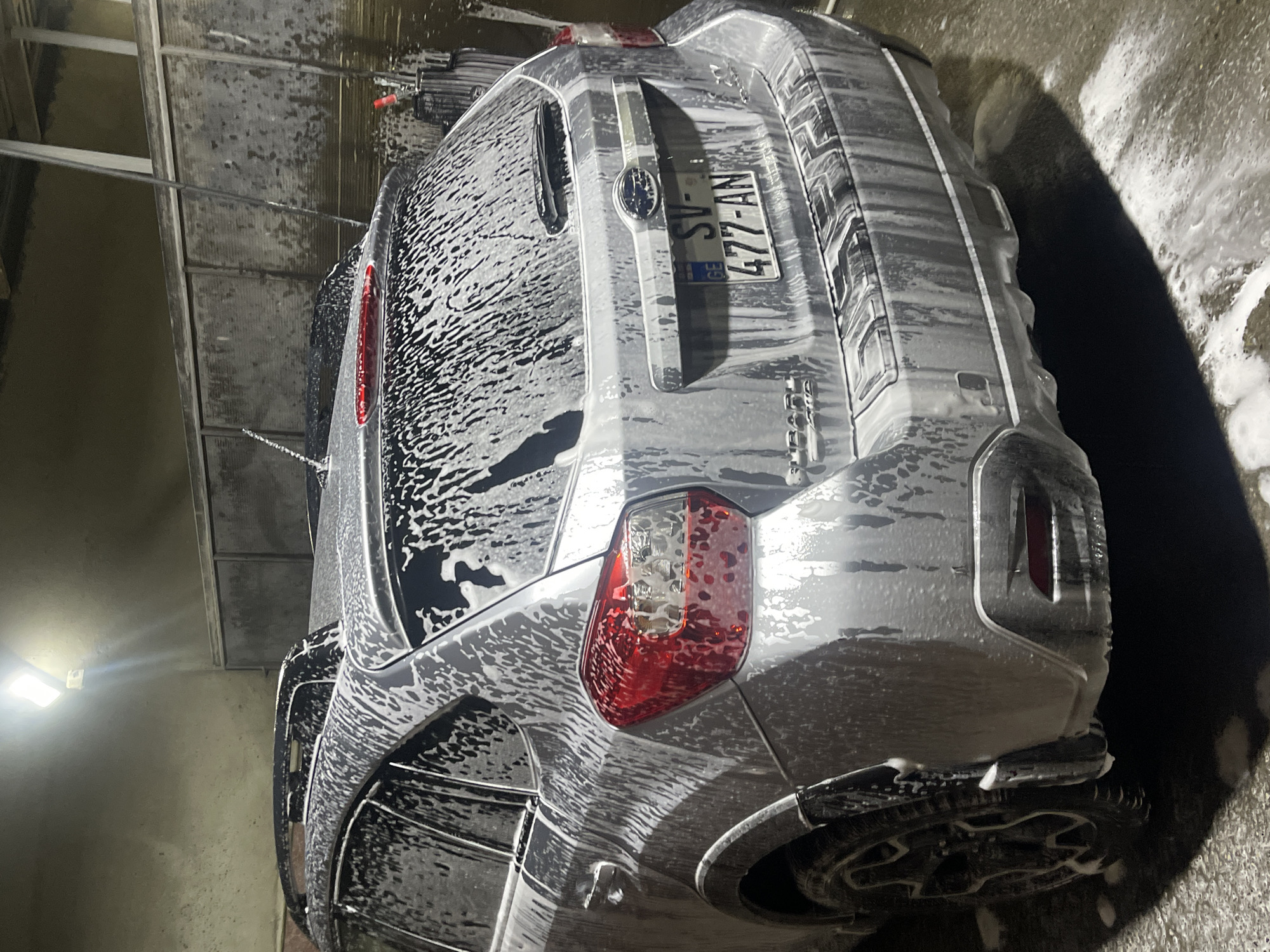 The image shows the rear of a silver Subaru vehicle undergoing a car wash. The vehicle is covered in white foam. The license plate reads "SV-477-AN" and indicates that the car is registered in Georgia. The letters "GE" are visible beneath the plate. The car is parked in what appears to be an enclosed car wash area, with structural elements of the car wash facility visible in the background. Based on the license plate and the location being specified as Tbilisi, Georgia, it can be inferred that this scene is situated in Tbilisi, Georgia.