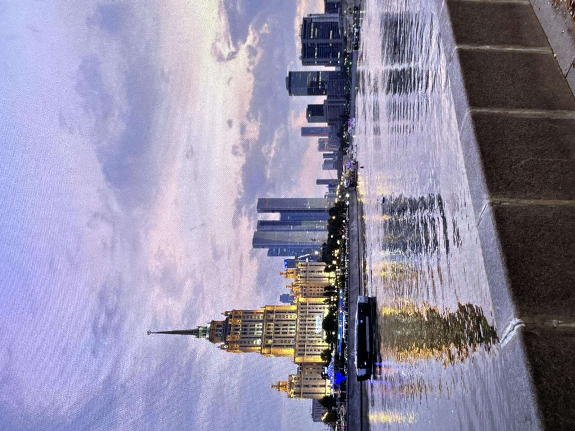 The image shows a waterfront view of a city skyline at dusk. A large, ornate, illuminated building with a spire is prominent on the left, with a series of modern skyscrapers to its right. A river or canal flows in the foreground, with a dark, sleek boat moving across it. The water reflects the lights of the buildings and the sky, which is a soft gradient of purple, pink, and blue with scattered clouds. A concrete embankment is visible in the bottom foreground. The overall scene suggests an urban environment at twilight, with an emphasis on architecture and the play of light. The location is identified as Pushkino, Russia.