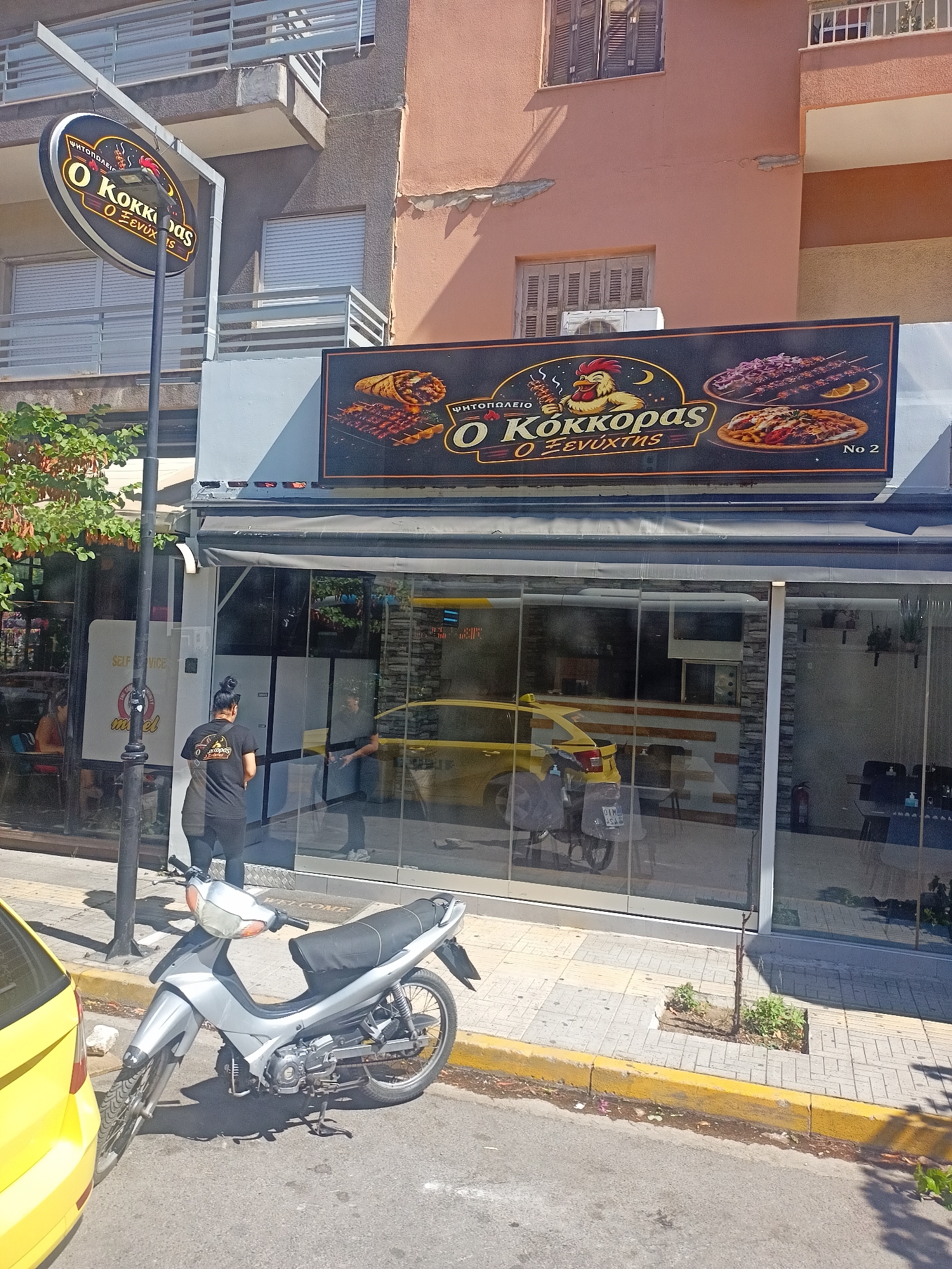 An outdoor, eye-level, slightly low-angle medium shot captures the facade of a restaurant called "O Kokkoras o Xenychtis" located in Unknown, Unknown. The restaurant features large, reflective glass windows and a black awning. Above the windows, a prominent black sign displays images of food items like souvlaki, gyros, and a salad, alongside the restaurant's name and logo, which includes a cartoon chicken. A circular, hanging sign with the same branding is visible to the left, supported by a metal pole.
In the foreground, a silver moped is parked on the sidewalk, partially obscured by a yellow vehicle on the left edge of the frame. The sidewalk is paved with grey and yellow tiles. A woman wearing a black t-shirt with the restaurant's logo and black leggings stands on the sidewalk, facing away from the camera and towards the restaurant entrance. A yellow car is visible through the glass windows of the restaurant.
The background shows the exterior of an apartment building with balconies and windows, some with wooden shutters. The overall scene depicts a street-level view of a food establishment on a sunny day.