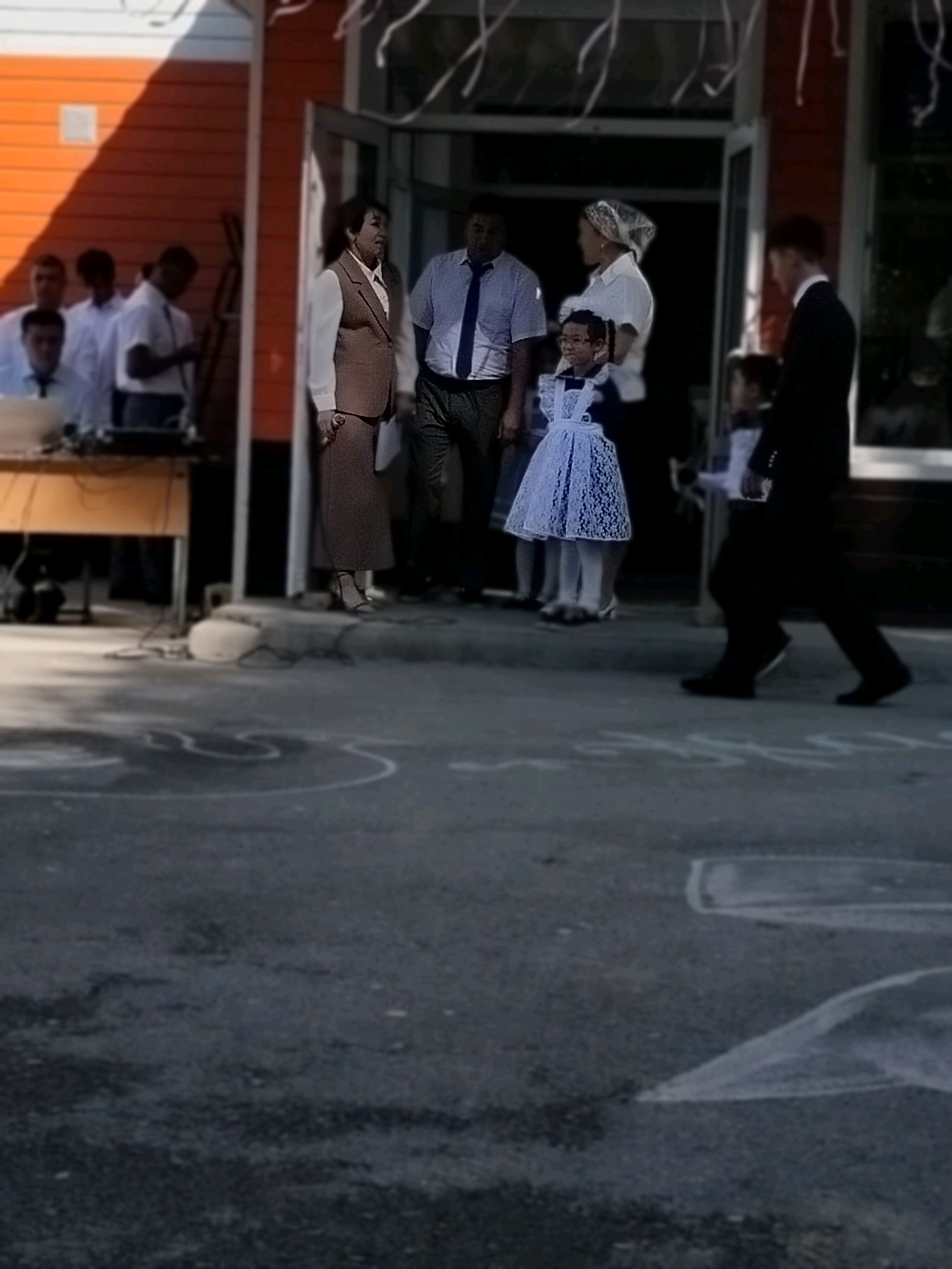 An outdoor scene in front of a building with an orange-colored wall and white trim. Several people are present. In the foreground, a young girl in a light blue, lacy dress stands with her hand to her mouth, looking to the right. Behind her, an adult woman in a brown suit and a man in a light blue shirt and dark tie are conversing. To the right of them, a man in a dark suit is walking away from the camera. Other individuals, some in white shirts and ties, are visible further back on the left side of the image. A wooden desk with equipment is situated on the left. The ground is paved with visible white markings, possibly indicating a social distancing or event layout. The overall impression is that of a gathering or event taking place outdoors.