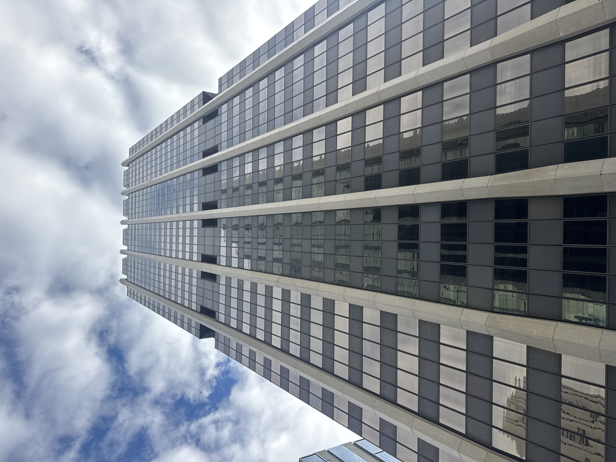 A low-angle shot of a tall skyscraper in Perth, Australia. The building's facade is composed of alternating bands of reflective glass windows and gray concrete. The windows reflect the sky and clouds, along with the reflection of a neighboring building. The sky features scattered clouds and patches of blue.