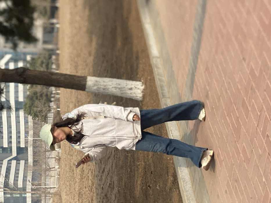 An individual, identified as a young female, is depicted walking on a reddish-brown brick paved pathway in the city of Zhongtun, China, during daylight hours. She is moving towards the left side of the frame, holding a light-colored smartphone in her right hand and appearing to view its screen. Her attire consists of a light-colored baseball cap, a light grey zippered hoodie featuring a dark logo on the left chest, blue wide-leg jeans, and light-colored athletic shoes. A light-colored backpack with light blue accents is worn over her shoulders. Her facial expression shows a slightly open or pursed mouth. To her right, a tree with a dark brown trunk, painted white at its base, stands next to the pathway. Beyond the path, the ground is covered in dry, light brown vegetation. In the background, several multi-story modern buildings are visible, interspersed with bare trees, under what appears to be a clear sky.