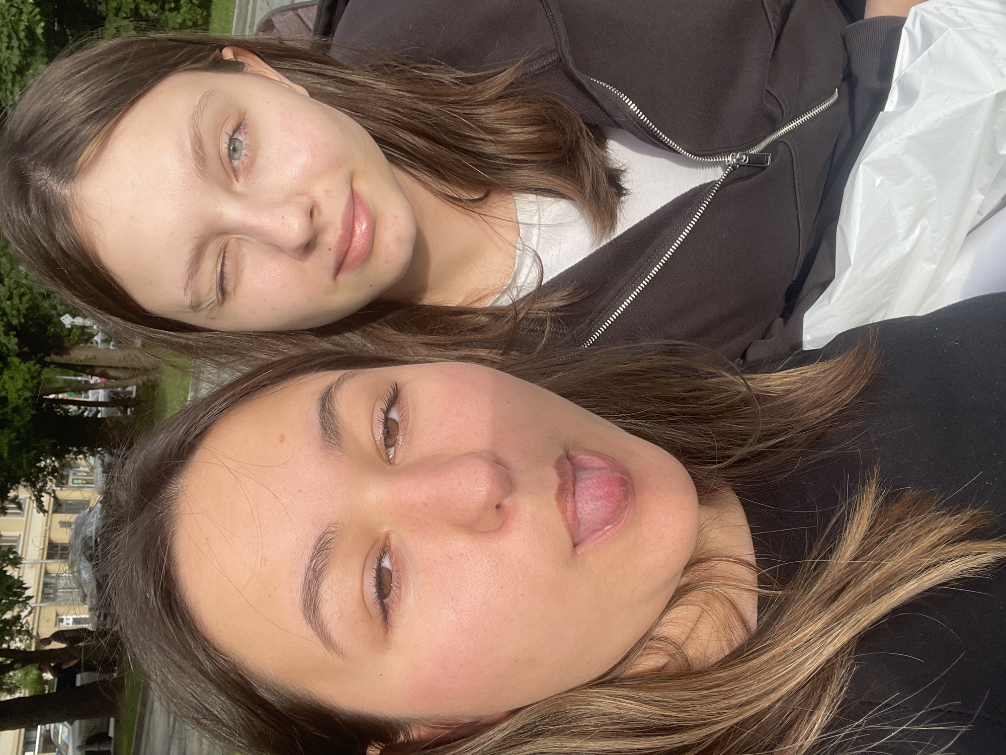 Two young women are captured in a close-up, low-angle selfie. The woman in the foreground is positioned on the bottom of the frame, with her face tilted upwards and her tongue playfully sticking out. Her skin is smooth and radiant, with defined eyebrows and warm brown eyes. Her hair, a mix of brown and blonde tones, is styled casually and falls around her face. She is wearing a black top.
Behind her and slightly above, the second woman is visible. She is winking, with one eye closed and the other open, looking towards the camera. Her lips are slightly parted, and she has a subtle smile. Her hair is also brown with lighter highlights, framing her face. She is wearing a dark brown zippered hoodie over a white t-shirt. A white object, possibly a bag, is partially visible next to her.
The background suggests an outdoor setting, with lush green trees and buildings that hint at an urban environment, consistent with Moscow, Russia. The lighting indicates it is daytime, likely with bright sunlight, casting a warm glow on the subjects' faces. The overall mood is lighthearted and casual, suggesting a moment of fun shared between friends. No text is visible in the image.