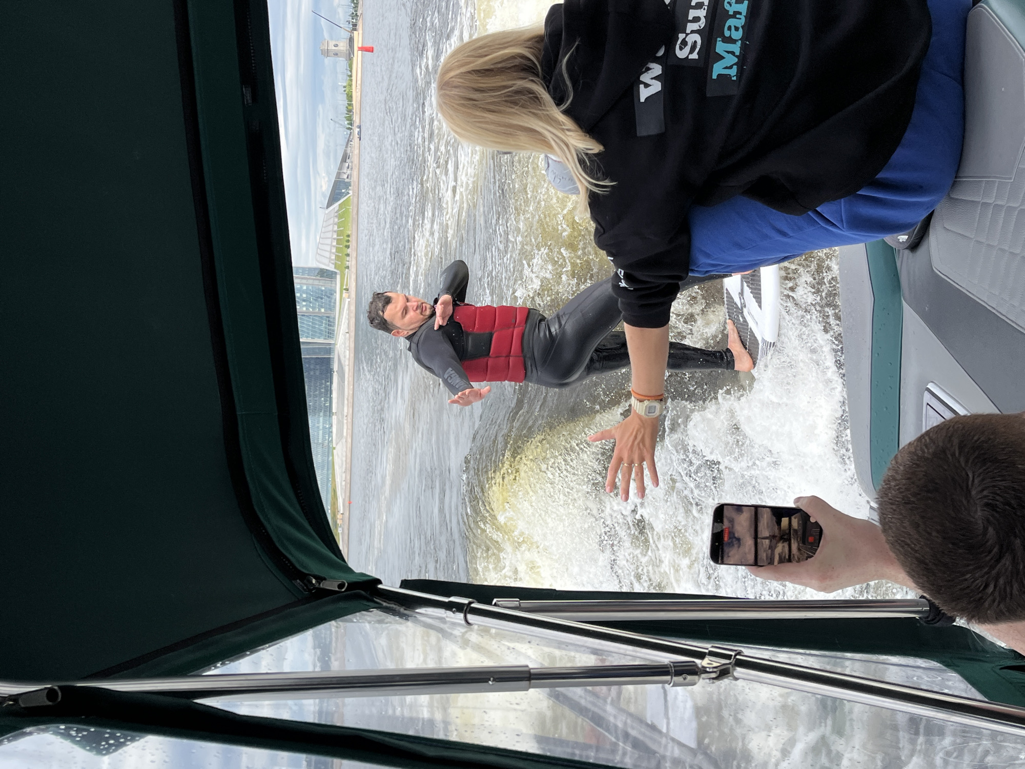 A man in a black wetsuit and a red life vest is wakeboarding in a river, with a woman in a black hoodie and blue sweatpants seated on a boat in the foreground. The man is in the water, riding a wakeboard and appearing to perform a trick. The woman is facing away from the camera, her hands extended. Another person is visible, holding a smartphone to record the scene. In the background, there are modern buildings and a cloudy sky, suggesting an urban setting. The water is choppy, with a significant wake created by the wakeboarder. The scene appears to be taking place on the Neva River in Saint Petersburg, Russia, given the architectural style of the buildings and the presence of a river in an urban environment.