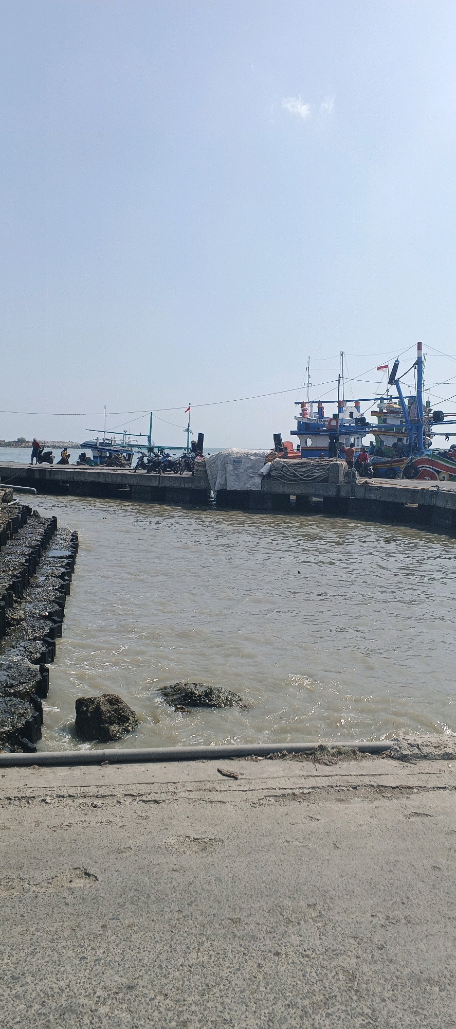 A wide shot shows a sunny day at a harbor in Blimbing, Indonesia. In the foreground, a concrete embankment runs horizontally across the bottom of the frame. To the left, a line of wooden posts topped with rocks forms a barrier in the murky water. The water itself is choppy and reflects the bright sky.
In the middle ground, a concrete pier extends into the water. Several fishing boats are docked along the pier. One boat, painted blue and white, has a red Indonesian flag flying from its mast. Another boat, larger and more colorful, is partially visible on the right side of the frame. Various people are seen on the pier, some standing and some seated. Several motorcycles are parked on the pier, and bundles of white material are covered with tarps on one of the boats.
The background features a hazy horizon where the sea meets a clear, pale blue sky. A few wisps of clouds are visible. The overall scene depicts a working harbor, likely a fishing community.