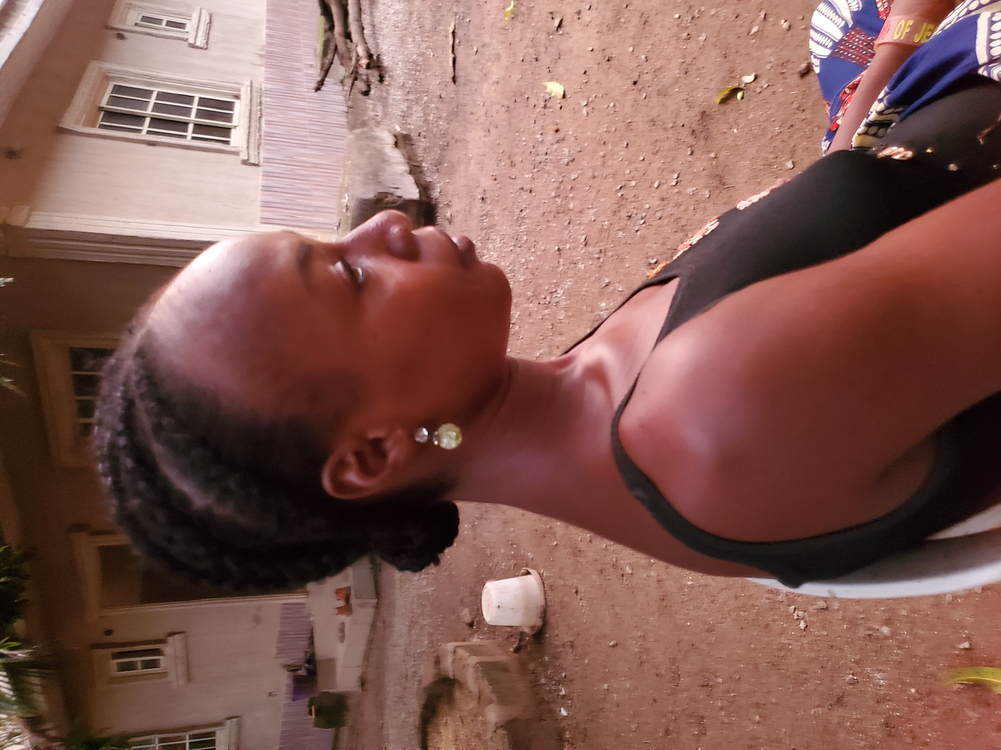 The image shows a dark-skinned woman with braided hair, wearing a black top, sitting outdoors. She is looking off-camera to the left. Behind her is a dirt ground, part of a building with white-framed windows, and a small white bucket. A portion of a patterned fabric is visible on the right side of the frame. The location is likely in Mararaba, Nigeria.
