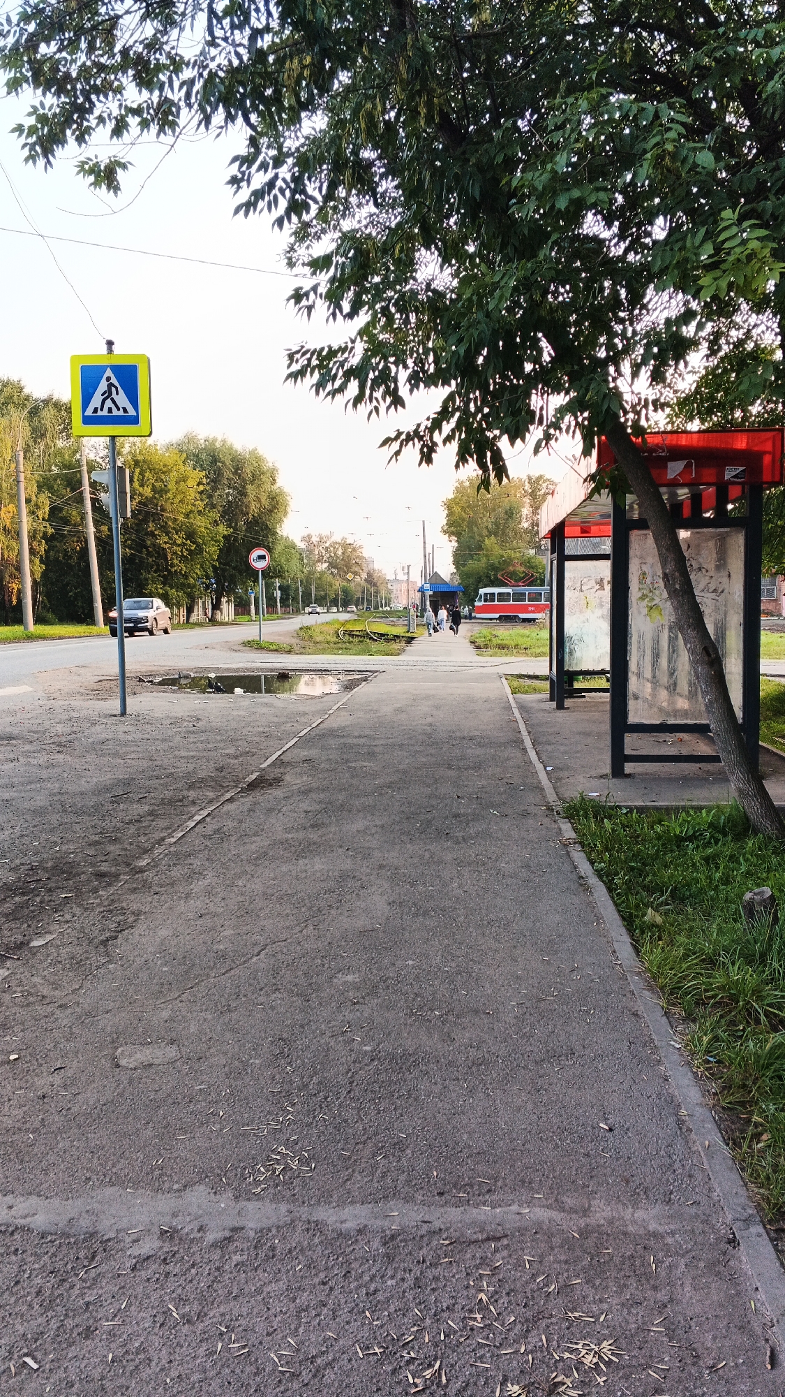 The image shows a street scene in Izhevsk, Russia. A pedestrian crossing sign is visible on the left side of the image. A bus stop shelter with a red roof is located on the right side. A red tram is seen in the distance. The sky is overcast, and the weather appears to be warm. The ground is paved with asphalt, and there is some grass and trees in the background. There are a few people walking on the sidewalk.