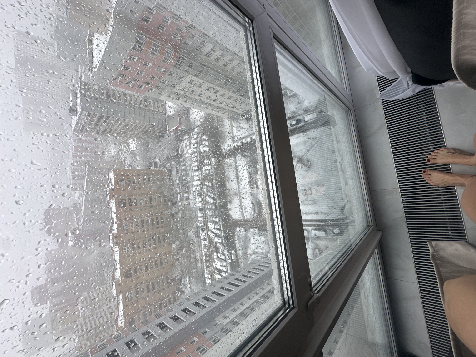 The image depicts a high-angle view from inside a building, looking out through a rain-streaked window. The external scene shows a dense urban environment, presumably in Moscow, Russia, characterized by multiple high-rise buildings with varying architectural styles. Streets and walkways are visible between buildings, suggesting a planned city layout. The weather appears overcast and rainy. Inside the building, a heating vent is visible beneath the window. A person's feet are partially visible near the edge of the frame. A white curtain and a beige towel are present near the window.