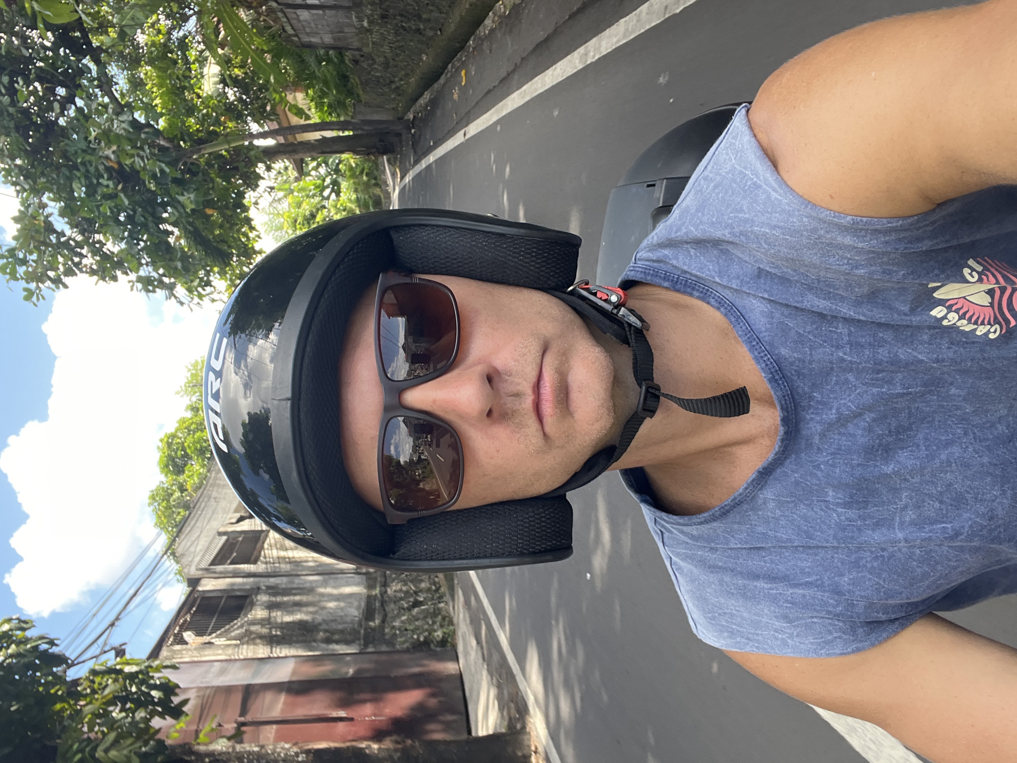 In North Kuta, Indonesia, a man wearing a black helmet and sunglasses rides a scooter. He is wearing a blue sleeveless t-shirt and is holding a phone up to take a selfie. The scooter is on a paved road with a white line marking the lane. On the left side of the road, there are buildings and trees. On the right side of the road, there are more trees and a wall. The sky is blue with some white clouds.