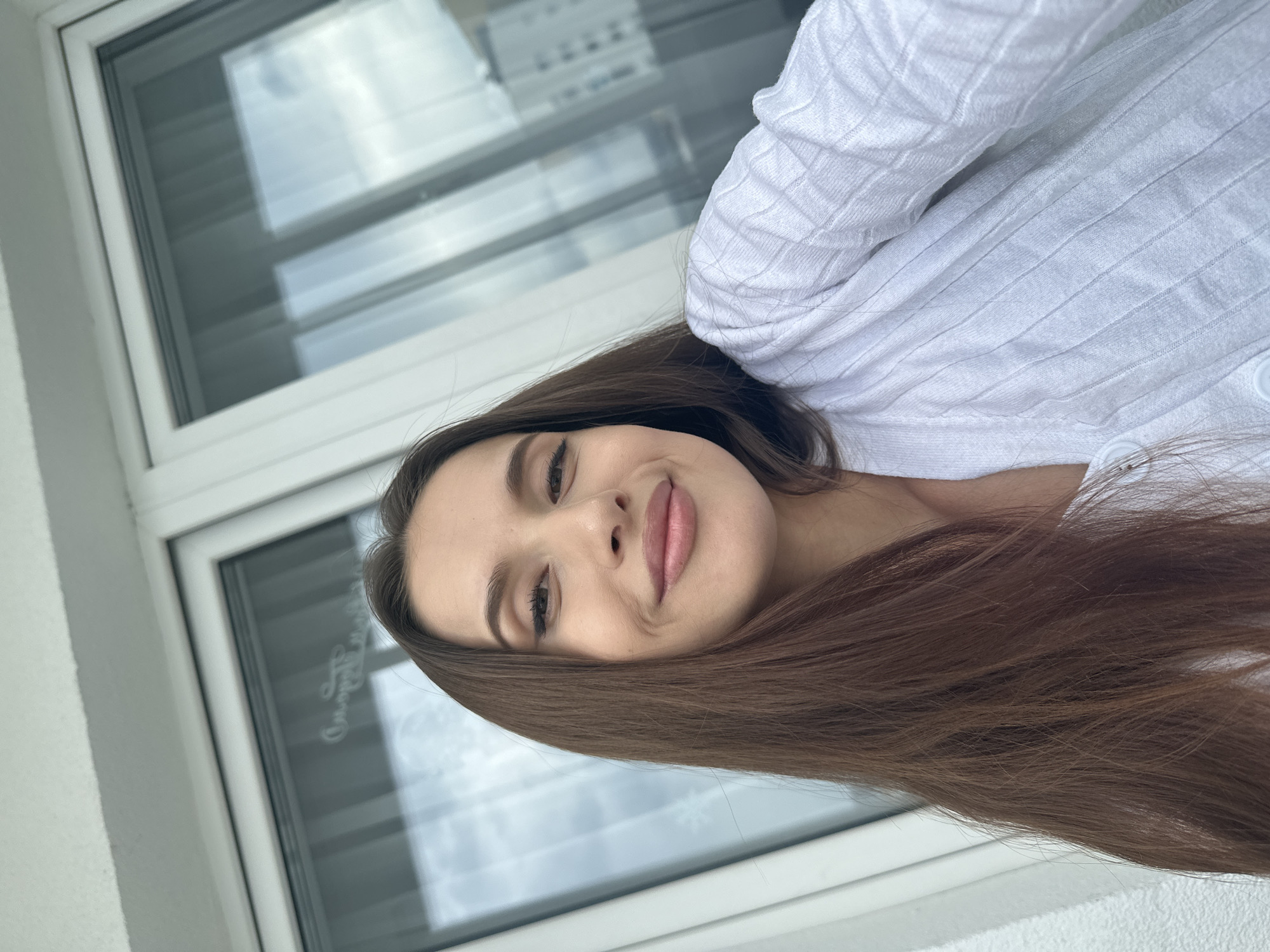 A young woman with long, straight, medium-brown hair and a subtle smile looks directly at the camera in a selfie. She wears a white, textured, ribbed cardigan with visible buttons. Her makeup features winged eyeliner and light pink lipstick. The background consists of large, white-framed windows reflecting an urban environment with distant buildings and a cloudy sky. A faint, decorative Cyrillic inscription, "Скоро Лето" (Soon Summer), is reflected on the glass of the window. The scene is located in the city of Yekaterinburg, Russia.