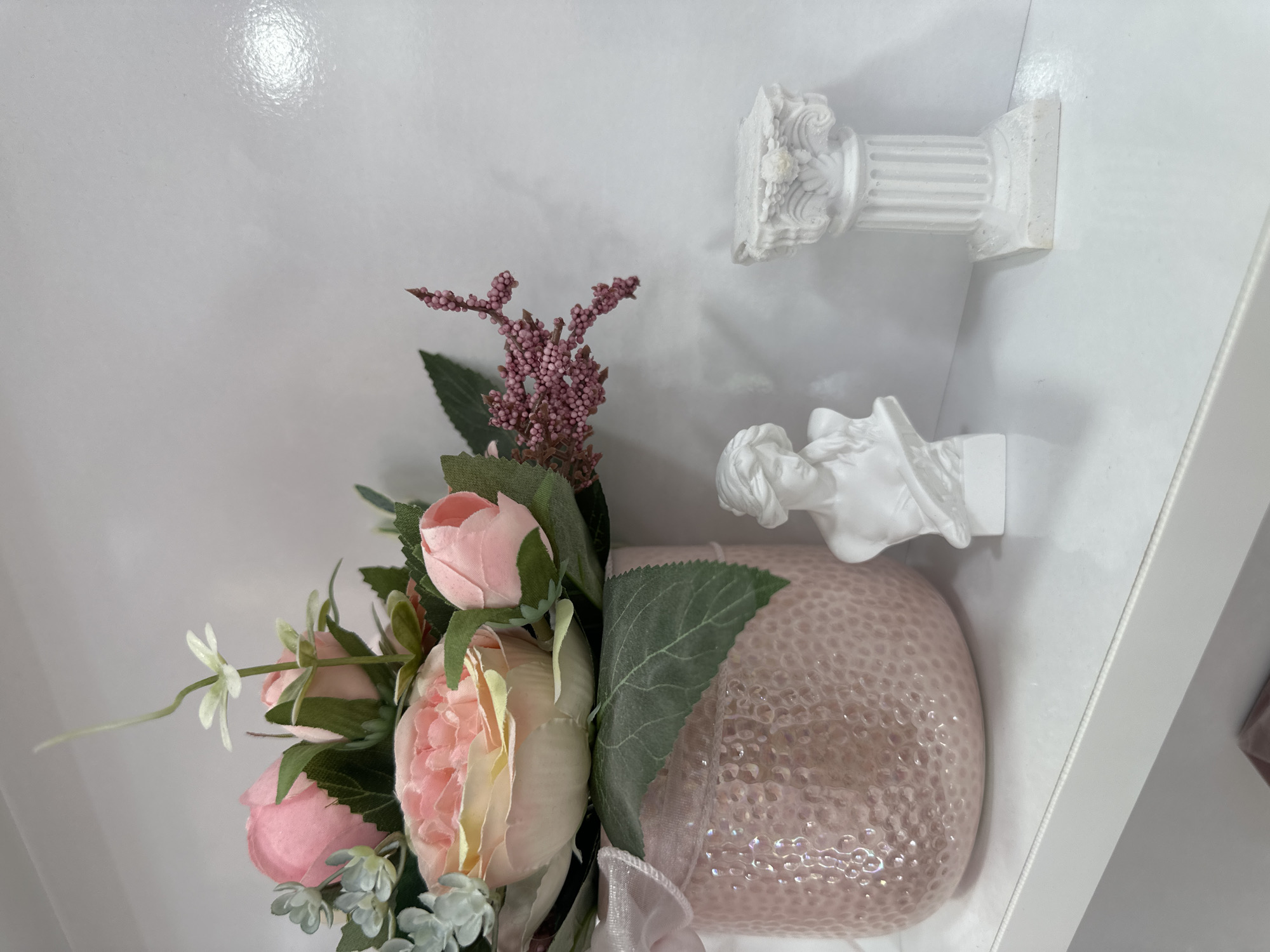 The image depicts a floral arrangement in a pink vase, accompanied by two small white sculptures. The objects are placed on a white shelf. There is no indication in the image of the location being Sofia, Bulgaria.