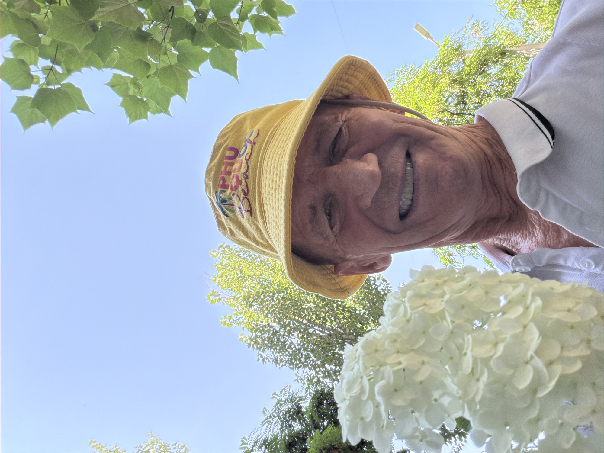 A low-angle, outdoor, medium close-up shot features a man wearing a yellow bucket hat with the text "PHU QUOC Beach" embroidered on it. He is also wearing a white polo shirt with black and white trim on the collar. The man is smiling and holding a large cluster of white hydrangea flowers in his left hand, which partially obscures his face. The background consists of a clear blue sky and the green leaves of trees, suggesting a bright, sunny day. The man's skin appears weathered, and he has wrinkles around his eyes and mouth. The overall impression is that of a man enjoying a summer day outdoors.
