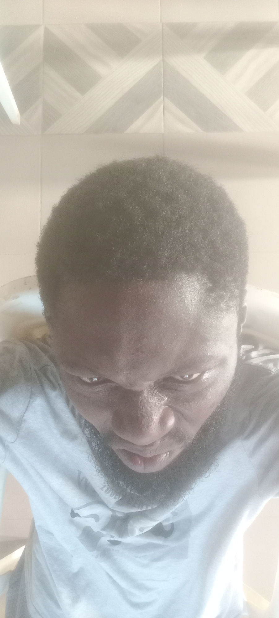A low-angle selfie depicts a dark-skinned man with short, dark hair and a beard. He is wearing a light-colored t-shirt. The background consists of tiled walls with a geometric pattern. The location is indeterminate, and the connection to Bende, Nigeria, is unsubstantiated from the visual data.