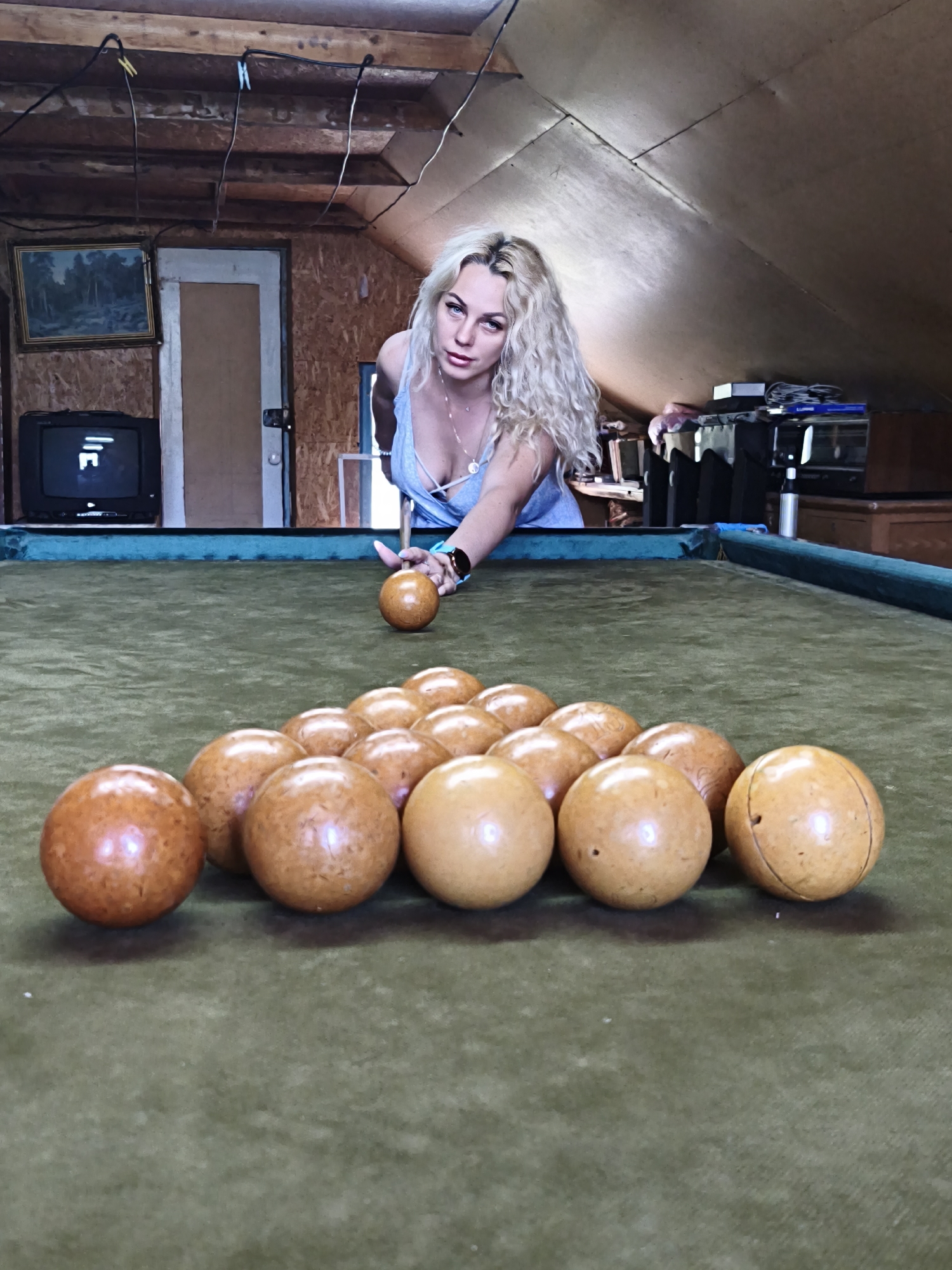 A woman with long, wavy blonde hair is in an attic or loft space in Терновский округ, Ukraine, playing on a billiard-style table. She wears a light blue, low-cut spaghetti strap top with multiple delicate necklaces and a watch on her left wrist. She leans over the green felt-covered table, holding a light brown ball in her right hand as if to strike it, while her left hand rests on the surface for stability. Her gaze is directed at the camera. Approximately 15 identical light brown balls are arranged in a triangular rack near the far end of the table. The table features blue-painted wooden rails. The environment is rustic: to the left, a black CRT television displays a white geometric pattern below a framed landscape painting. A wooden door is partially visible behind these, set into an unfinished plywood wall. The ceiling consists of exposed wooden beams with black electrical wires strung across them and secured by clothespins. To the right, the wall slopes sharply, with various objects including boxes and cables on a surface. The walls and ceiling are constructed from visible wooden panels and plywood.