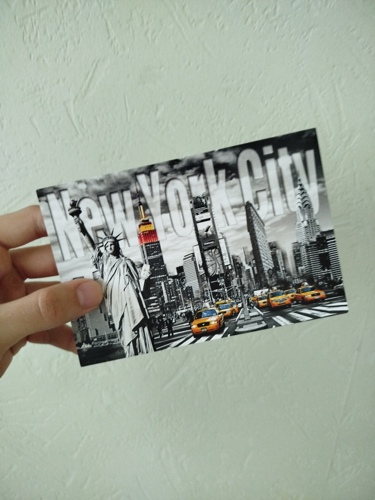 A hand holds a postcard featuring a black-and-white image of New York City. The image includes the Statue of Liberty, the Empire State Building, Times Square, and yellow taxis. The text "New York City" is superimposed over the skyline. The background behind the postcard is a textured, off-white wall. The location is not Varna, Bulgaria.