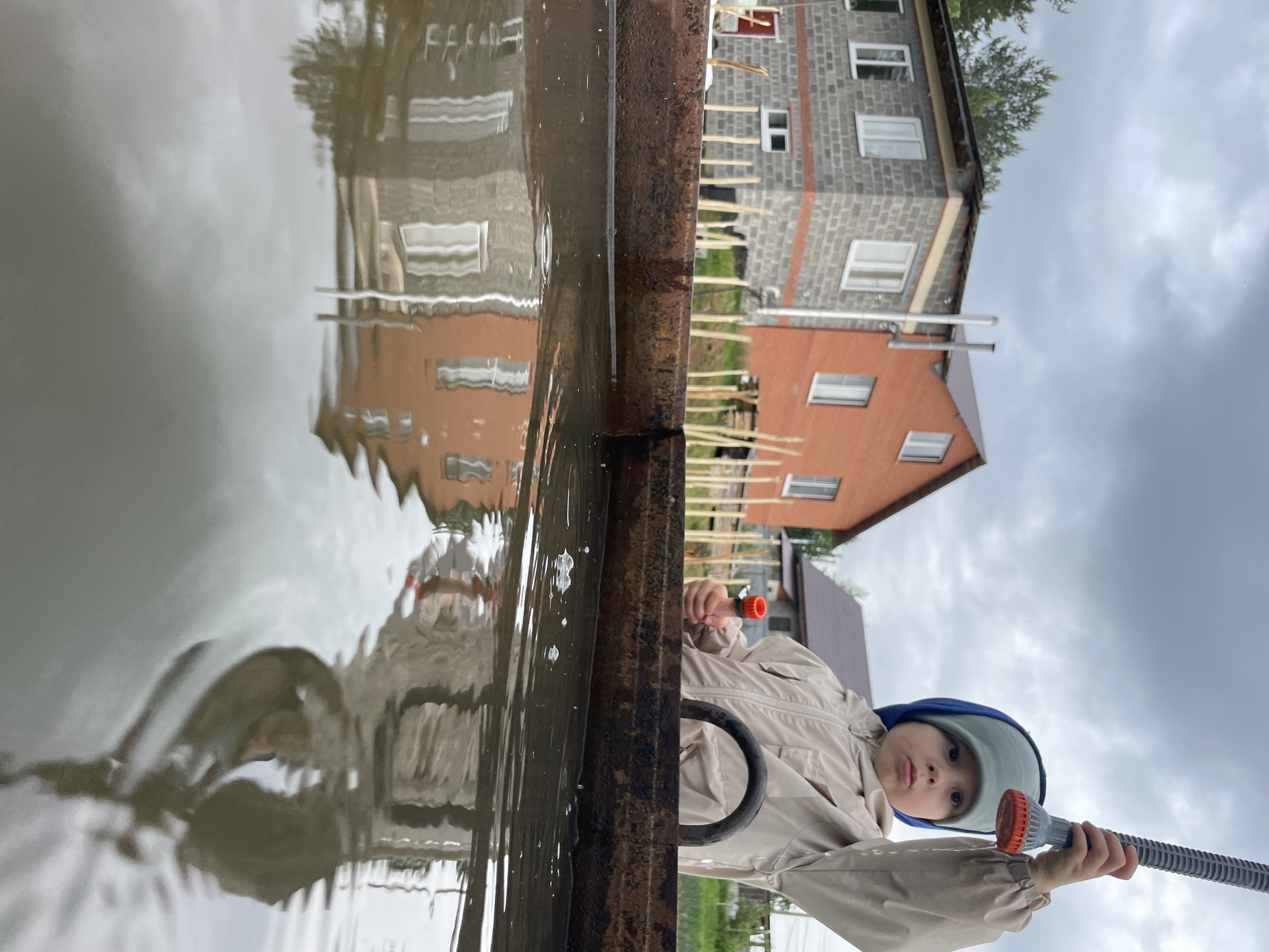 The image depicts a low-angle view from a body of murky water within a rusty, dark metal trough, looking up at a young child and residential structures under an overcast sky. A toddler, wearing a light-colored jumpsuit and a light blue/grey hat, stands behind the trough, holding a grey ridged hose nozzle with an orange end in their raised right hand. Their left hand holds a smaller orange object. Part of a black, circular object is visible in front of the child.
In the midground, two houses are visible. The house on the left is a two-story structure made of grey concrete blocks with white-framed windows and orange-red horizontal banding, appearing to be under construction with a partially finished roof. To its right, a two-story red brick house with white-framed windows and a dark grey pitched roof features a metal chimney pipe on its side. Between the trough and the houses, a line of numerous thin, light-colored wooden stakes or poles is driven into the ground. Green coniferous trees are visible behind the grey house, suggesting a wooded area. The sky is filled with dark grey clouds. Reflections of the houses and the child are visible in the rippled water, along with small bubbles on the surface. The scene is outdoors, likely in a suburban or rural area undergoing development.