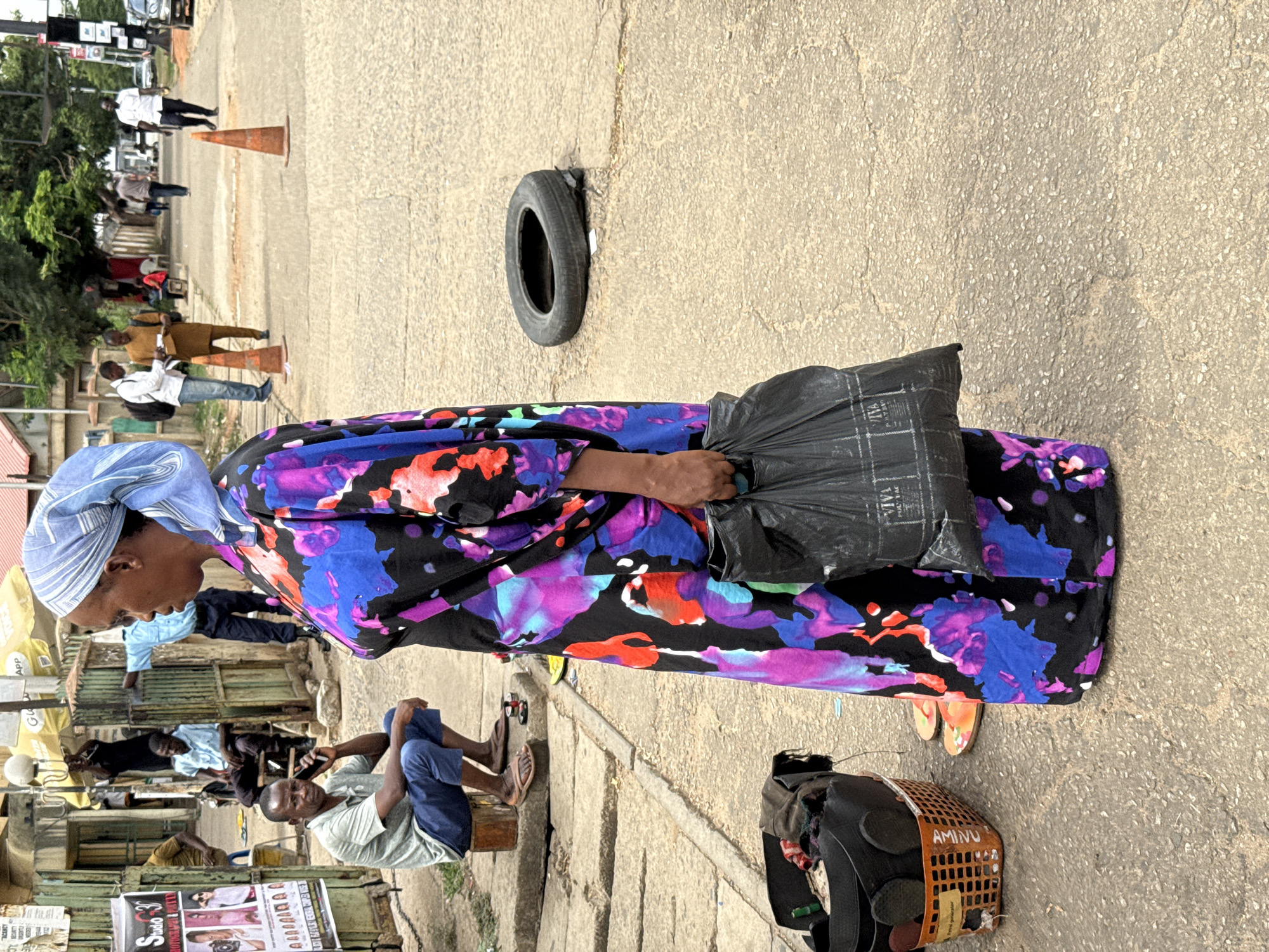 A medium-shot photograph depicts a woman in a vibrant blue, purple, and black patterned dress, carrying a black plastic bag. She wears a pale blue headscarf and is standing on a paved street next to a raised, segmented sidewalk.
On the sidewalk, a man is sitting on a block using his phone. The scene contains multiple vendors and various subjects.
Lying on the street near the woman is a car tire. Further down the street, road cones, and additional pedestrians are visible.
The scene is presumed to be set in Abuja, Nigeria.