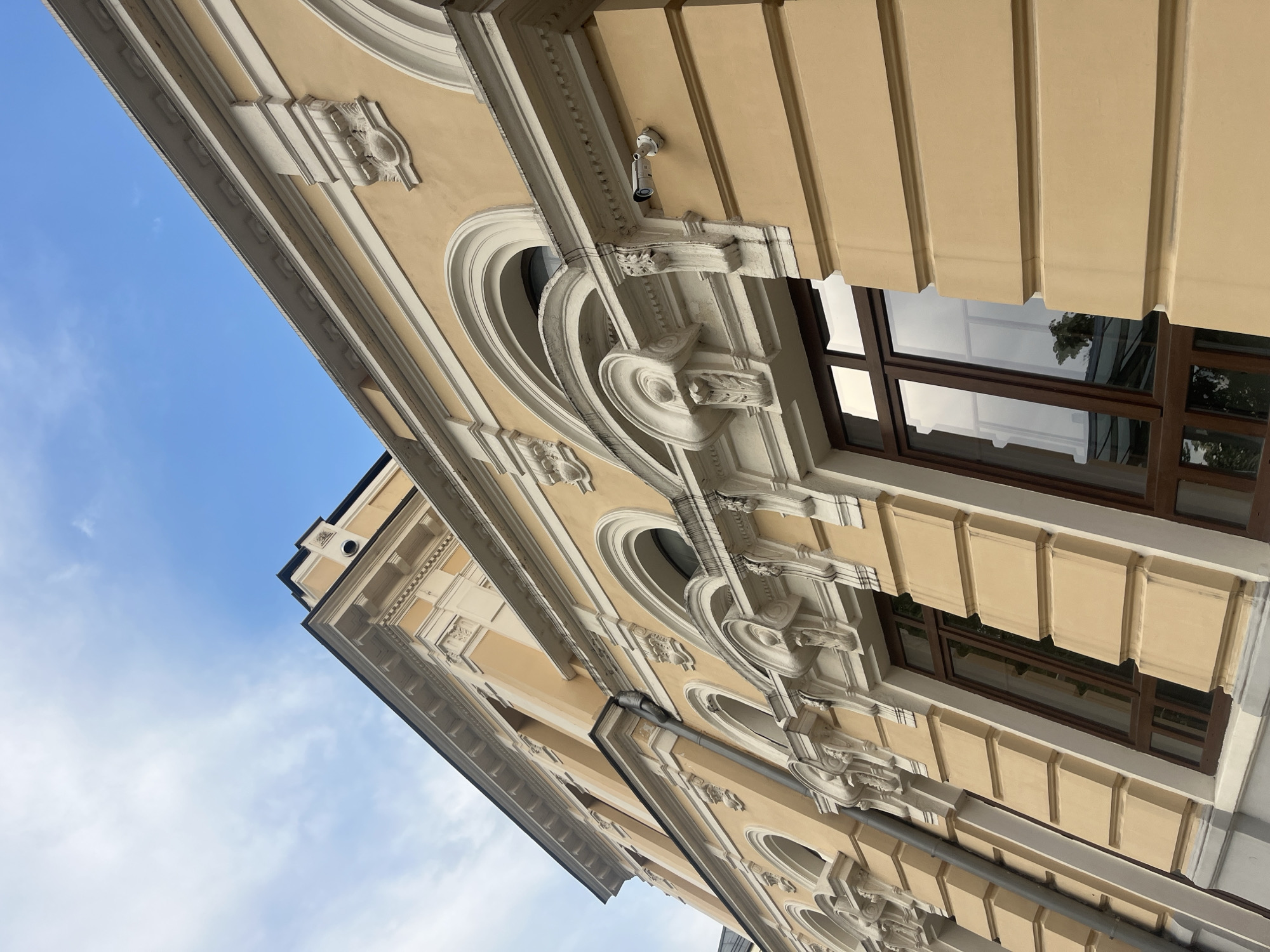 This is a low-angle shot of the facade of a building in Plovdiv, Bulgaria. The building is painted in a pale yellow color and features ornate white trim around its arched windows. Some of the windows are visible, with dark brown frames. A security camera is mounted on the facade. The sky above is blue with some white clouds. The image appears to be taken during the daytime.