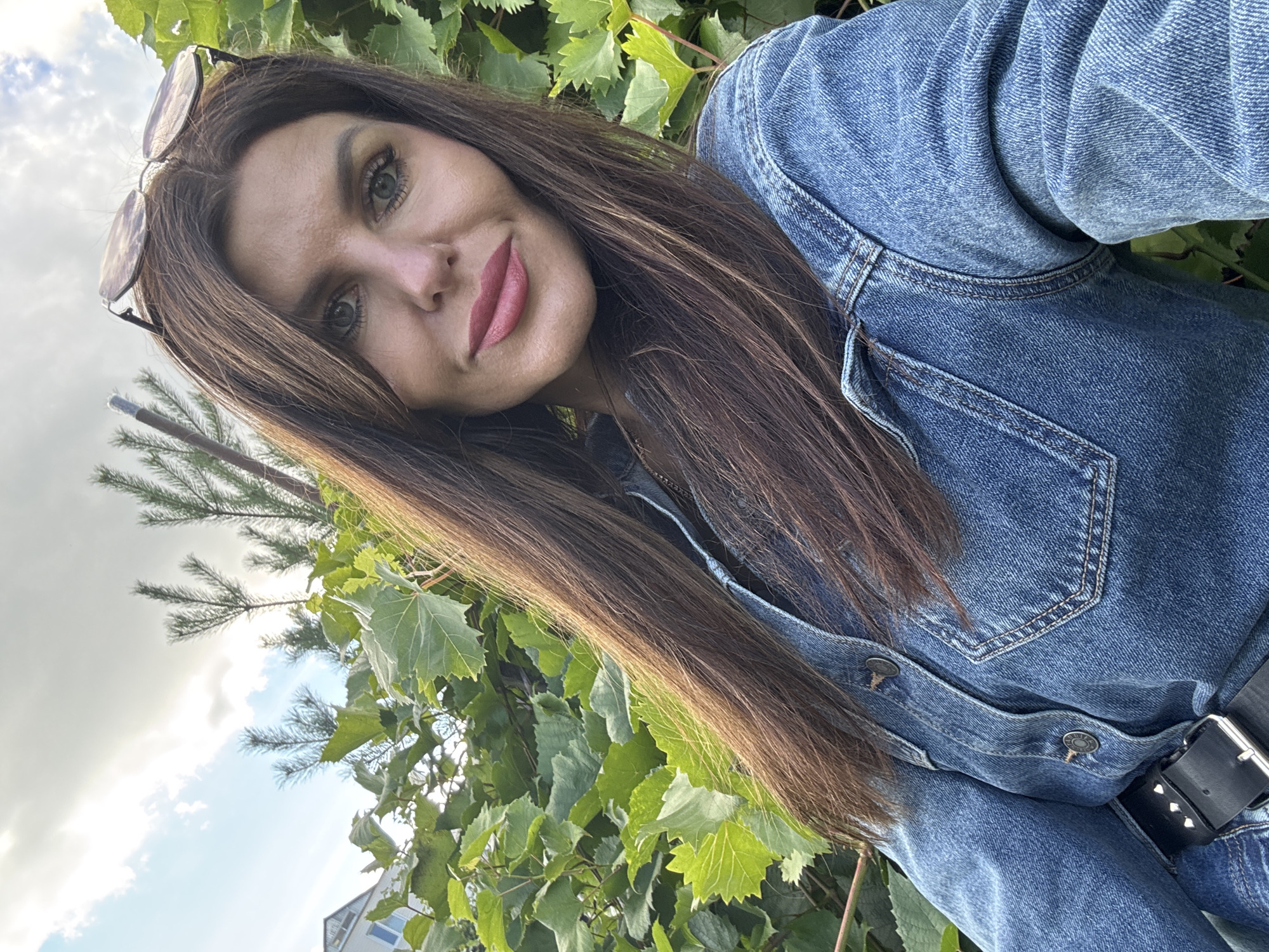 In a medium shot, a woman with long, brown hair and blue eyes looks into the camera, wearing a denim jacket and sunglasses perched on her head. She is outdoors, with green foliage behind her and a cloudy sky above. The scene appears to be in a garden or park, possibly in Dubovaya, Russia, given the presence of what might be a building and trees in the background. The lighting suggests it is daytime, with the sun partially obscured by clouds. The woman's expression is neutral, and she is not interacting with anyone else in the frame.