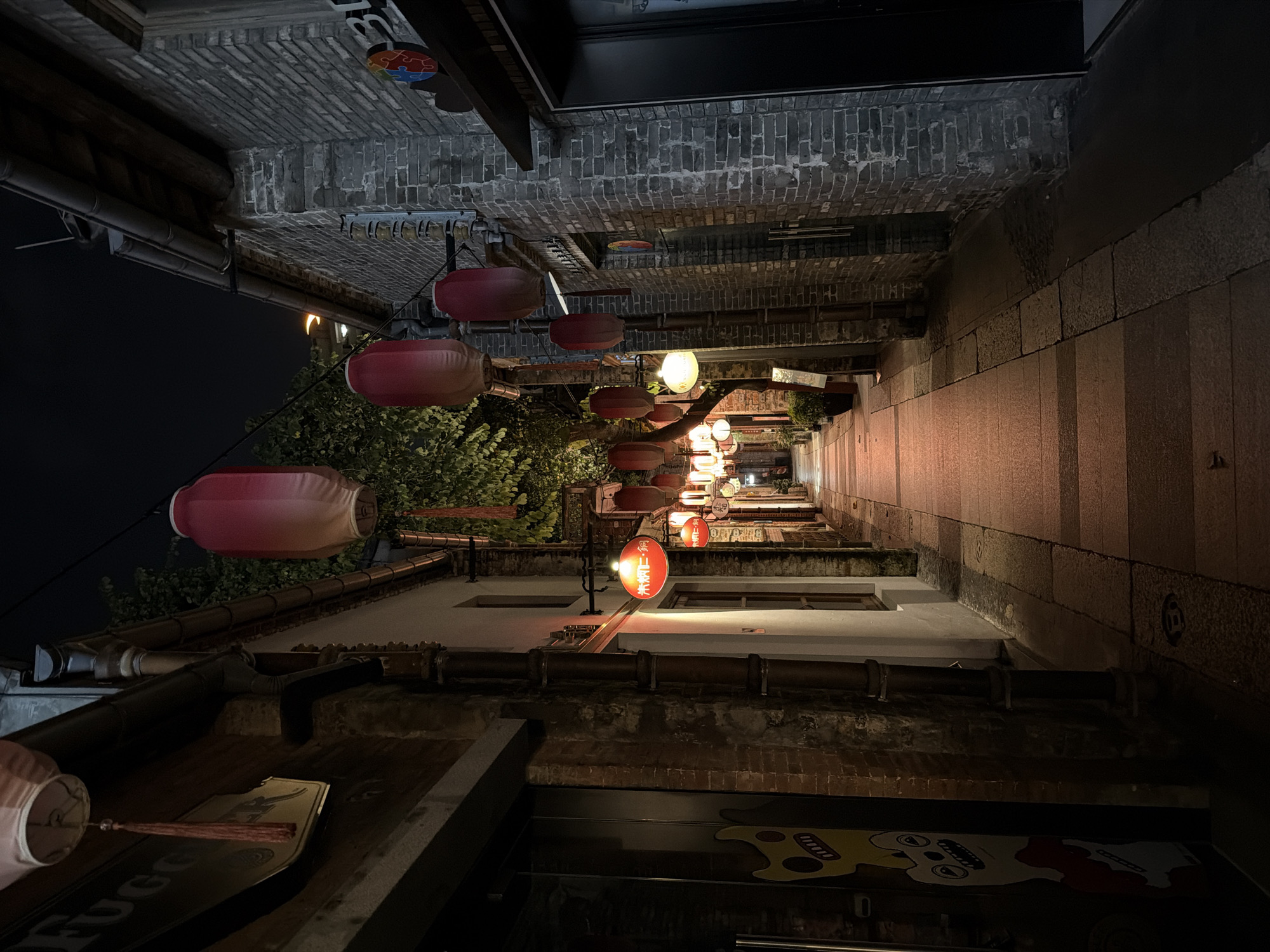 The image depicts a narrow alleyway at night in Chancheng District, China. The alley is lined with traditional brick buildings on both sides. Red and pink paper lanterns are strung across the alley, casting a warm glow on the stone-paved walkway. Lush green trees with visible foliage are present, adding a touch of nature to the urban setting. The buildings feature architectural details such as wooden doors and windows, and some have visible signage. One building on the left displays a cartoonish, yellow and white graphic. The overall atmosphere is serene and atmospheric, suggesting a quiet evening in a historic part of the city.
