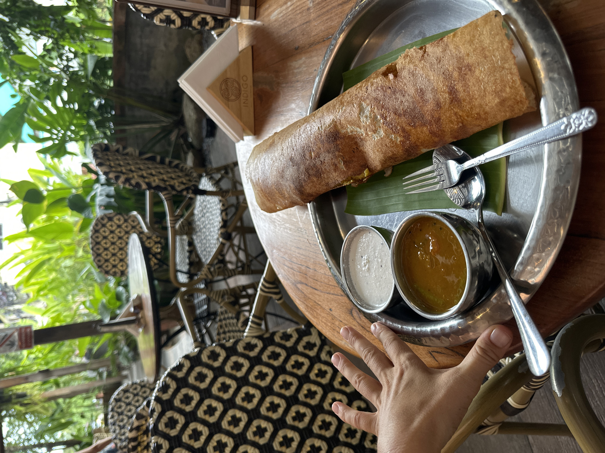 A hand reaches into the frame from the left, near two small metal bowls containing a white condiment and an orange curry, placed on a round metal plate. A long, golden-brown dosa is centered on the plate, resting on a green banana leaf. A fork and spoon rest on the plate beside the dosa. The plate is on a wooden table, with a patterned chair and blurred greenery visible in the background. A napkin dispenser with the logo "INDIGO" is present on the table. This image was taken in Koh Phangan, Thailand.