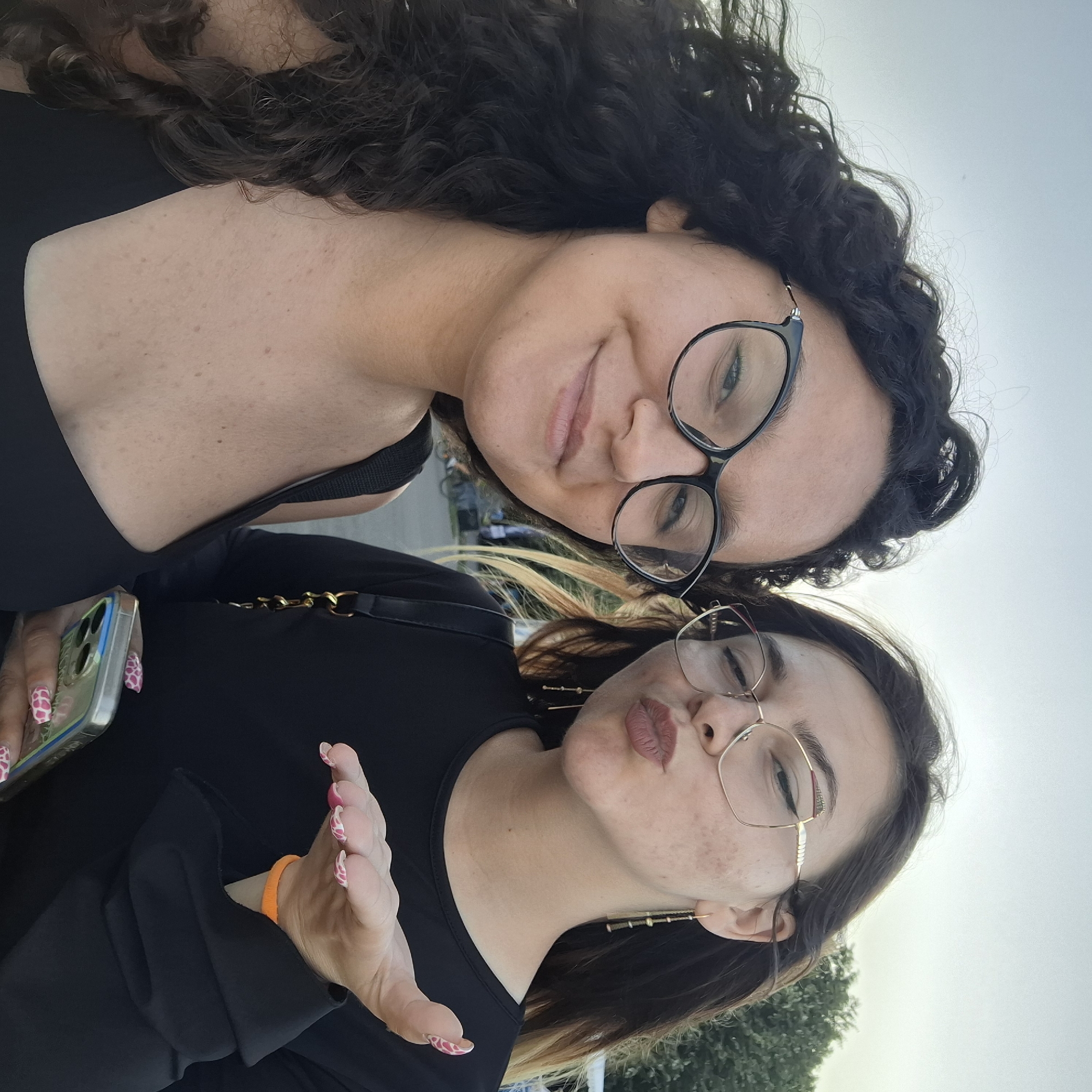 Two women are posing for a selfie outdoors in an urban environment. The woman on the left has dark, curly hair and is wearing glasses. She is smiling at the camera. The woman on the right is wearing glasses and is puckering her lips as if to blow a kiss, and extends her hand towards the camera. She has on a black top and a gold chain purse. In the background, trees and a street are visible. The sky is overcast.
