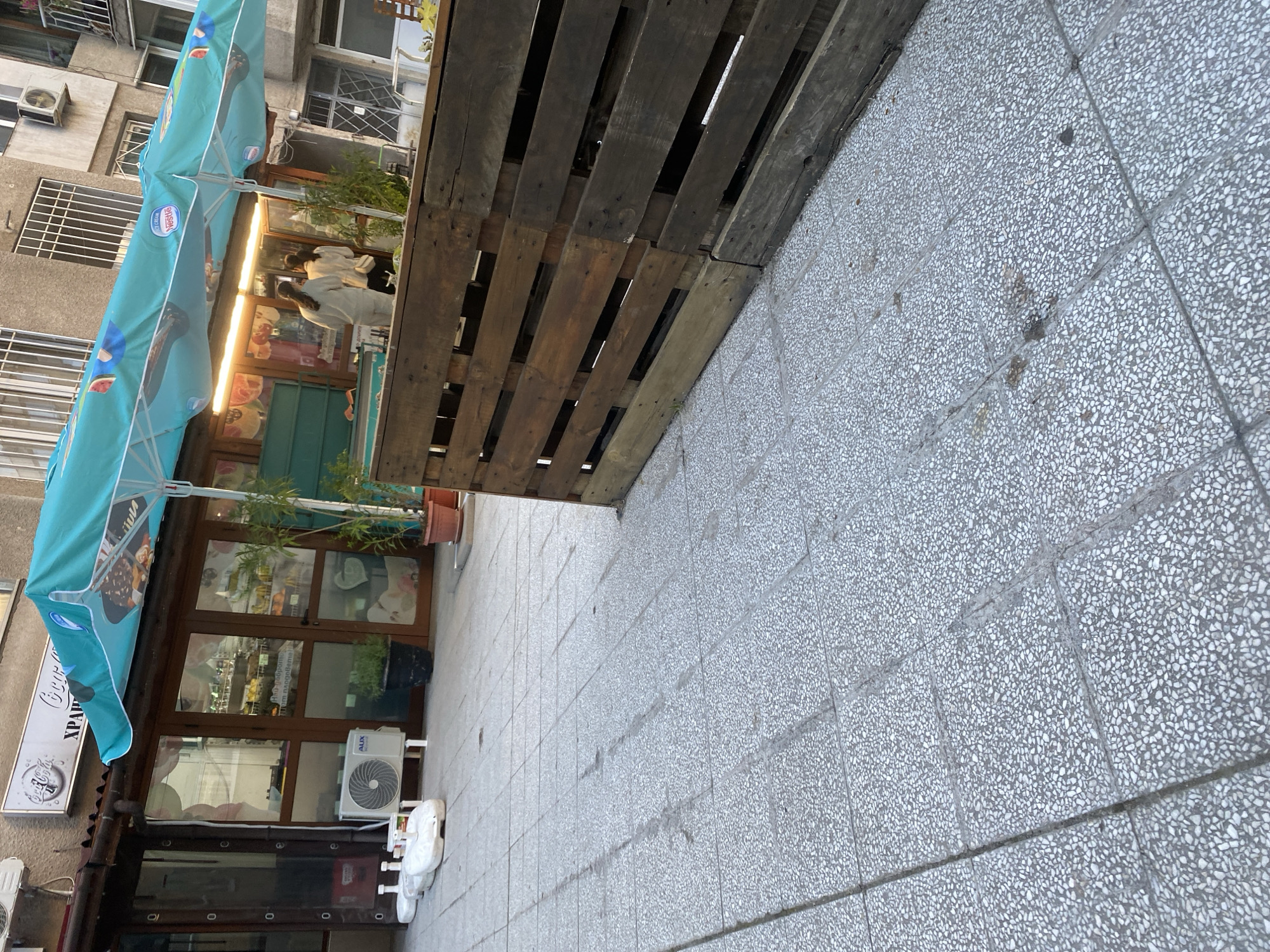 The image depicts the exterior of a shop in Sofia, Bulgaria. A wooden pallet serves as a temporary barrier or decorative element along the sidewalk. A blue parasol stands in front of the shop, partially obscuring the facade. The shop's storefront features large glass windows and a door, with a Coca-Cola sign visible on the left side of the door. The building is typical of urban construction in Sofia, with multiple floors. The sidewalk is paved with gray tiles. Two people wearing white clothes are standing near the display case of the shop. An air conditioning unit is mounted on the wall below the shop's front window. Several white plastic chairs are stacked on the sidewalk.