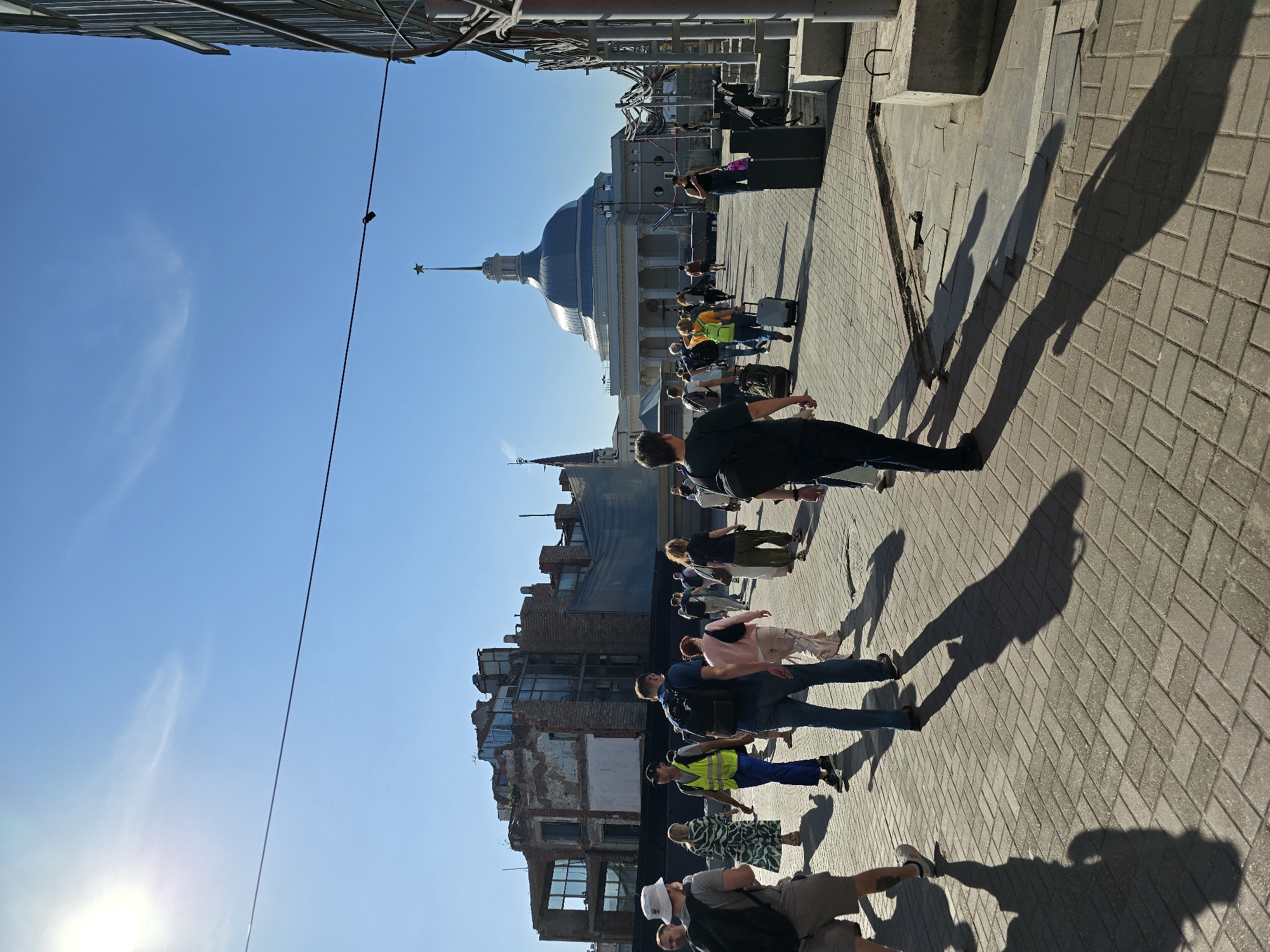The image depicts a sunny day in Moscow, Russia, likely near a transportation hub, given the presence of people with luggage and the architectural style. In the foreground, a group of people are walking on a paved walkway. The individuals are dressed in casual attire, with some carrying backpacks and rolling suitcases. The sun casts long shadows on the ground, indicating it is either morning or late afternoon.
In the background, a distinctive domed building with a spire topped by a star is visible. This building is likely a landmark of some significance, possibly a train station or a public building. To the left of this structure, there is a partially demolished or under renovation building with exposed brickwork and broken windows, suggesting ongoing construction or urban renewal. The sky is clear and blue, with a faint wispy cloud. On the right side of the image, a modern building with a metallic facade is partially visible, contrasting with the older architecture. The overall scene conveys a sense of bustling urban activity on a clear day.