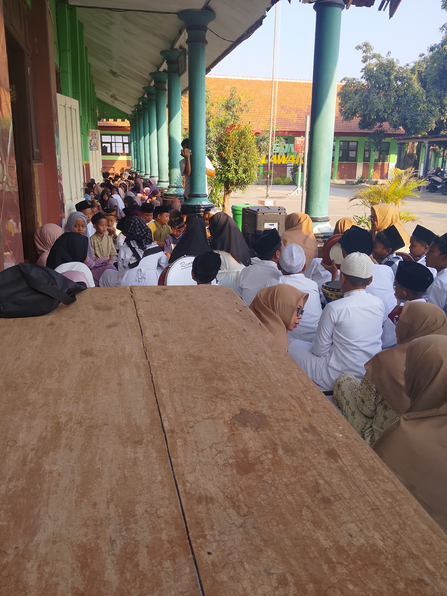 In Kepuh, Indonesia, a group of children and young people are gathered outdoors, likely at a school or community event. They are sitting on the ground, some on a wooden bench or platform in the foreground, and others further back under a covered walkway with green pillars. Many of the individuals are wearing white clothing and black or white skullcaps, suggesting a religious or cultural occasion. Some are wearing hijabs. In the background, a building with an orange tiled roof and green trim is visible, along with trees and a clear blue sky. One person in the distance appears to be holding a phone. A drum is visible among the seated individuals.