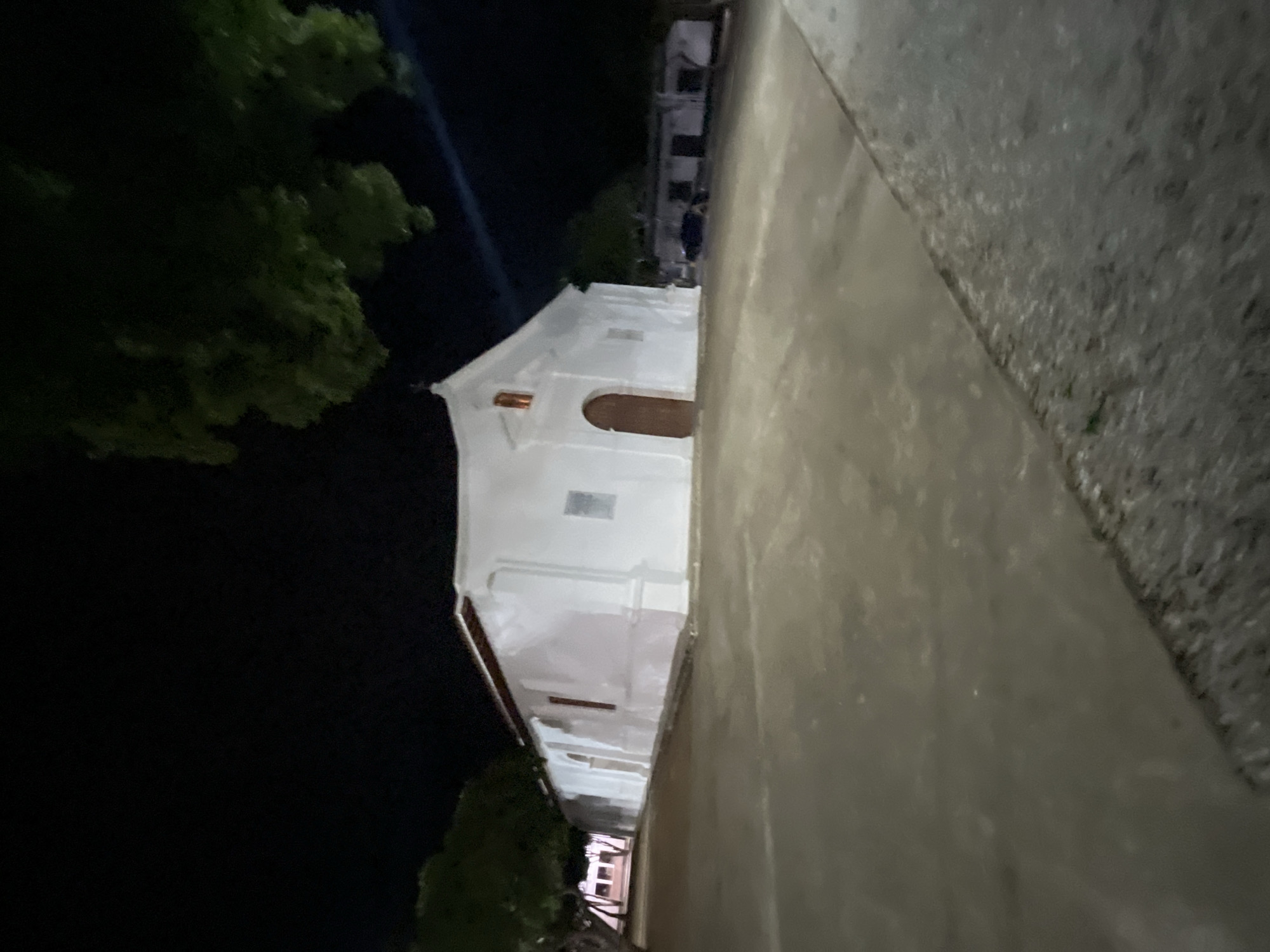 The image shows a white church at night. The church has a single wooden door and a small, lit window above it. There are trees on the left side of the image and a dark sky above. The ground in front of the church is paved. In the background, there are other white buildings with dark roofs. A dark-colored vehicle is parked in front of these buildings. The scene is located in the city of Santa Ana, Venezuela.