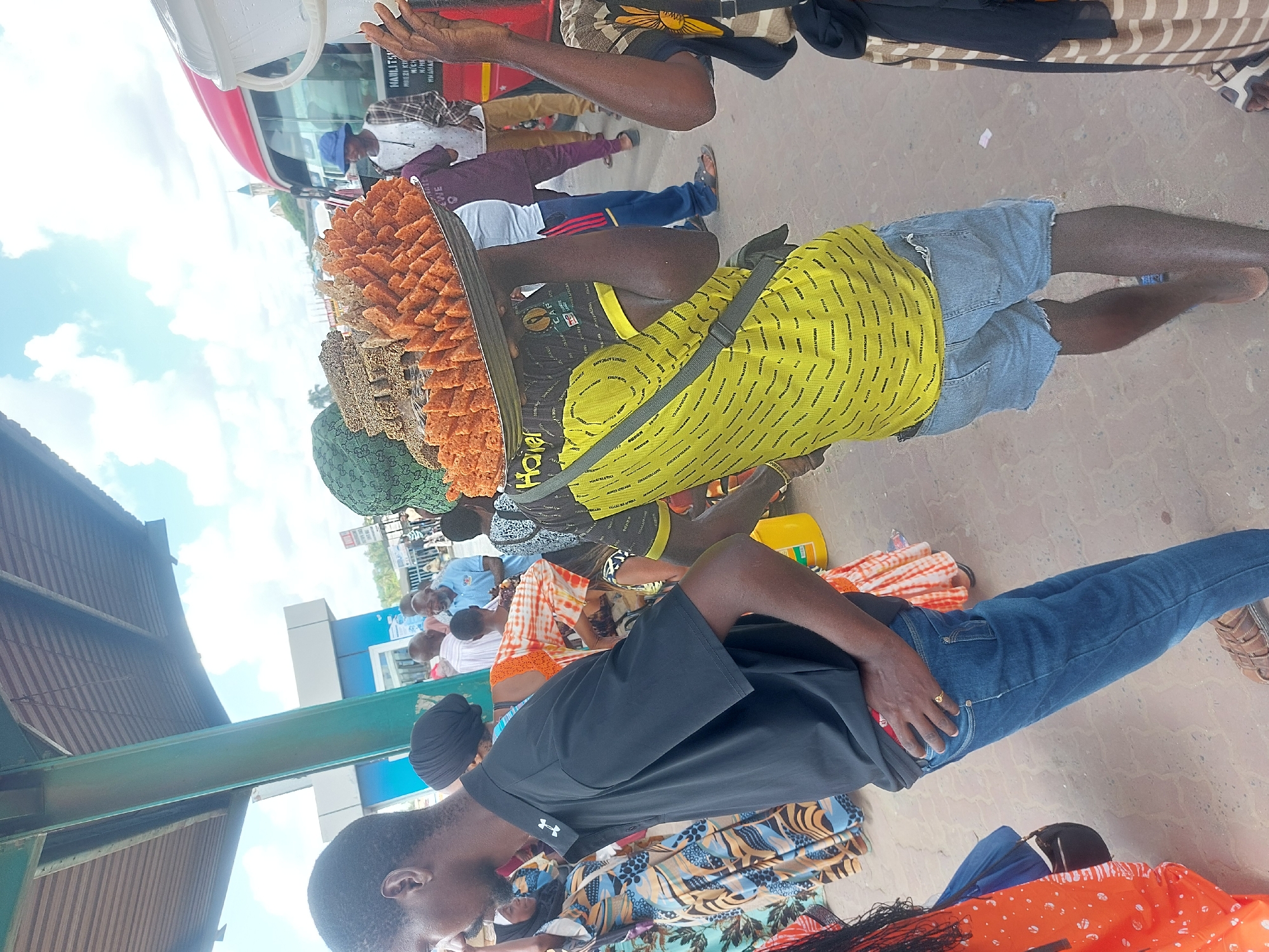 The image shows a street scene, probably in Dar es-Salaam, Tanzania, given the context. A man is carrying a large tray of what appears to be fried snacks or pastries, possibly a type of local delicacy. He wears a yellow shirt with dark text, blue denim shorts, and a satchel strap across his chest. Other people are visible in the background, some sitting, some standing. There is a bus or vehicle in the background, and the presence of other individuals suggests a public area or marketplace. The environment looks like a busy commercial or transportation hub. The photo has been taken at a slight angle to the right.