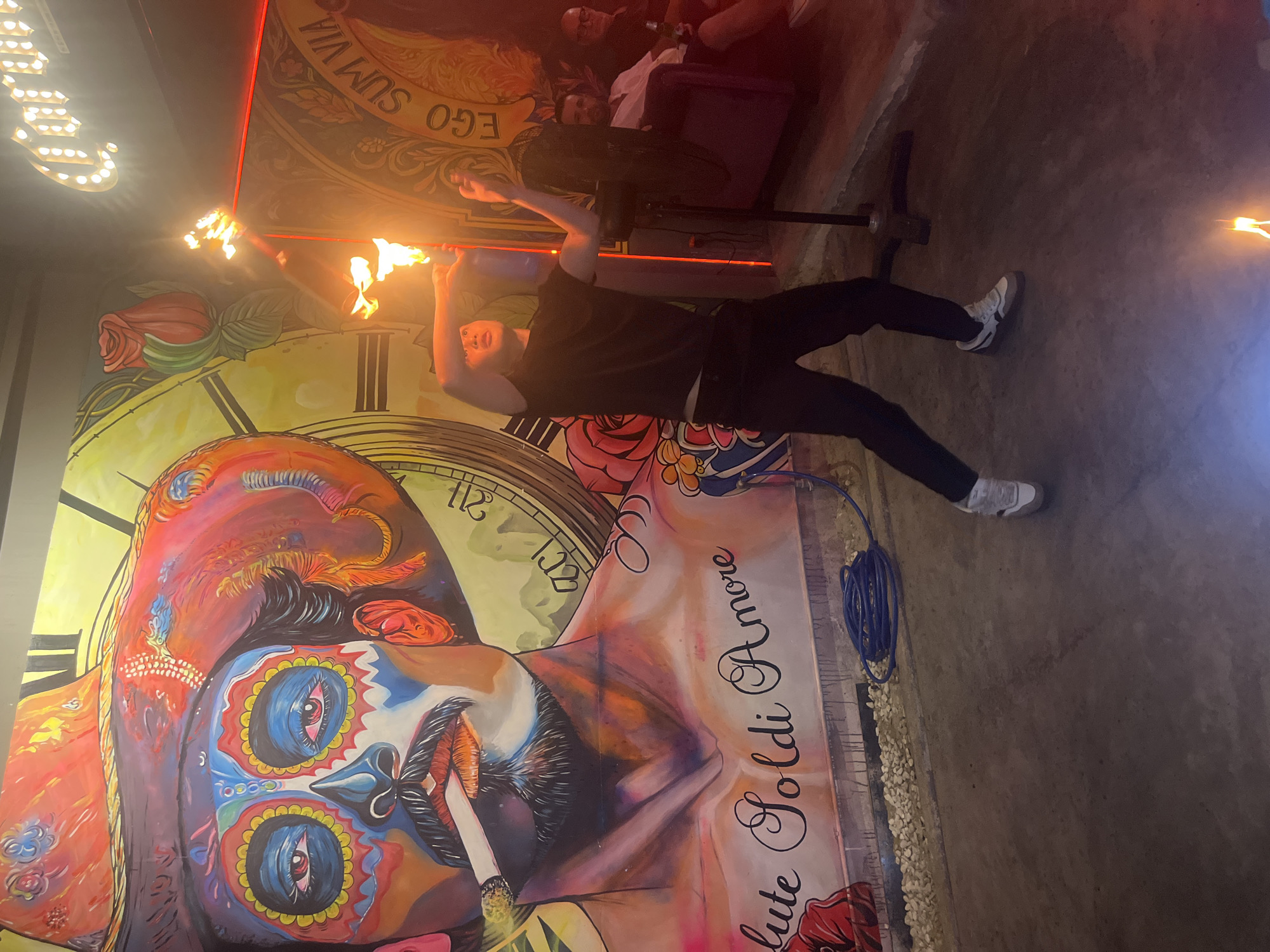 In Da Nang, Vietnam, a performer is captured mid-action at night, holding a flaming torch aloft. The backdrop is a vibrant mural featuring a stylized portrait of a person with face paint resembling a Day of the Dead skull, wearing a large, colorful hat, and smoking a cigarette. The mural also includes a large clock face and floral designs. The performer, dressed in black pants and a black t-shirt, stands on a concrete floor, with a fire visible on the ground near their feet. To the right, three men are seated on a purple couch, with one holding a bottle. The environment appears to be an indoor entertainment venue.