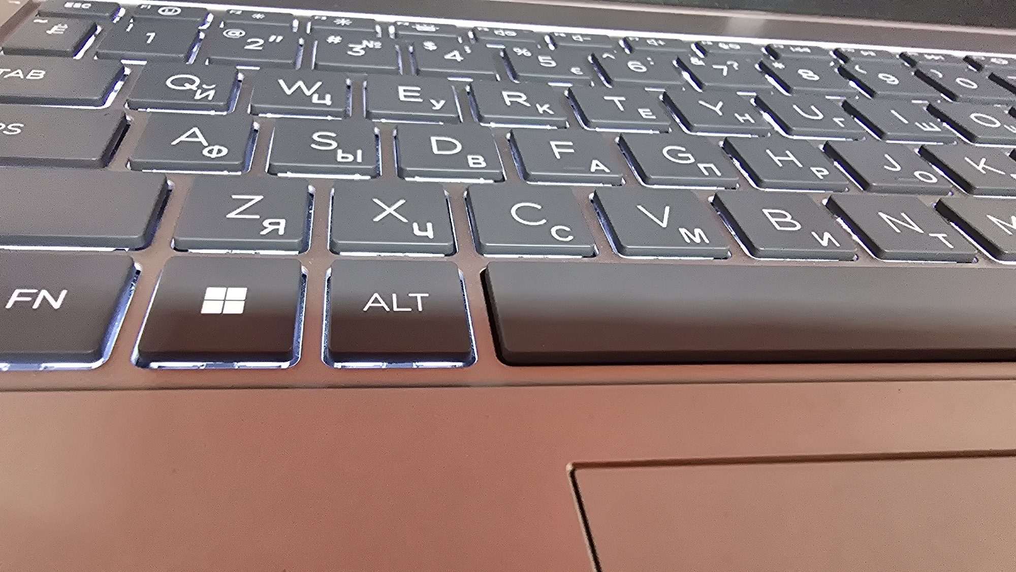 A close-up, low-angle shot shows a portion of a backlit keyboard. The keys are dark gray with white lettering, illuminated by a cool blue light from beneath. The layout suggests a Cyrillic keyboard, as indicated by characters like "Ф", "Ы", "Я", "Ц", and "У". Visible keys include "FN", a Windows logo key, "ALT", and various letter keys. The keyboard appears to be on a laptop, with the right side of the image showing a section of the laptop's body in a warm, reddish-brown hue. The surface of the laptop body is matte. The background is out of focus, suggesting the image was taken indoors. There are no people or animals visible in the image.
